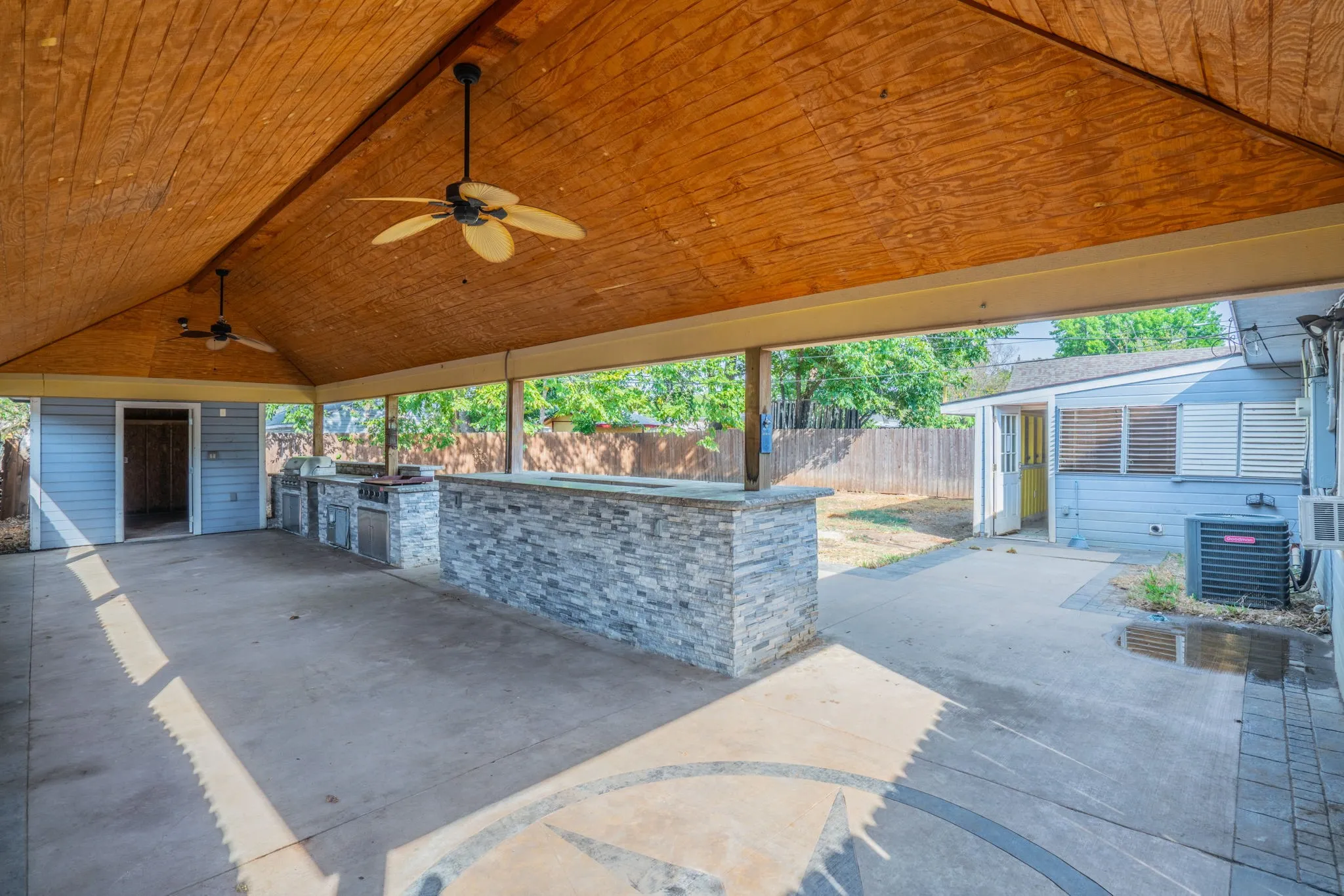 Expansive covered patio featuring a wood-finish ceiling, dual ceiling fans, and an outdoor kitchen with integrated appliances and a stone-clad bar
