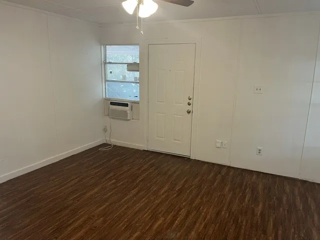 Wood-finish flooring, white paneled walls, ceiling fan, window-mounted air conditioning unit, and a solid white door