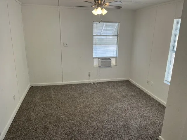Neutral wall paneling, gray carpet flooring, window with blinds, ceiling fan with light fixture, wall-mounted air conditioning unit