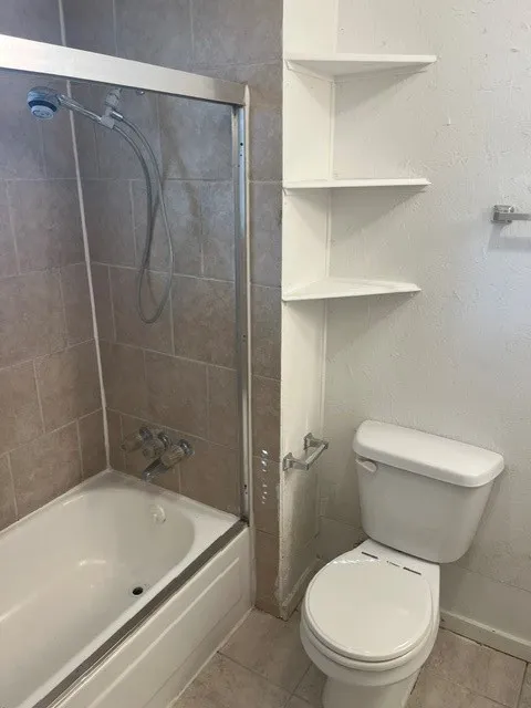 Bathtub and shower combination with a sliding glass enclosure, tile surround, white toilet, and built-in corner shelving
