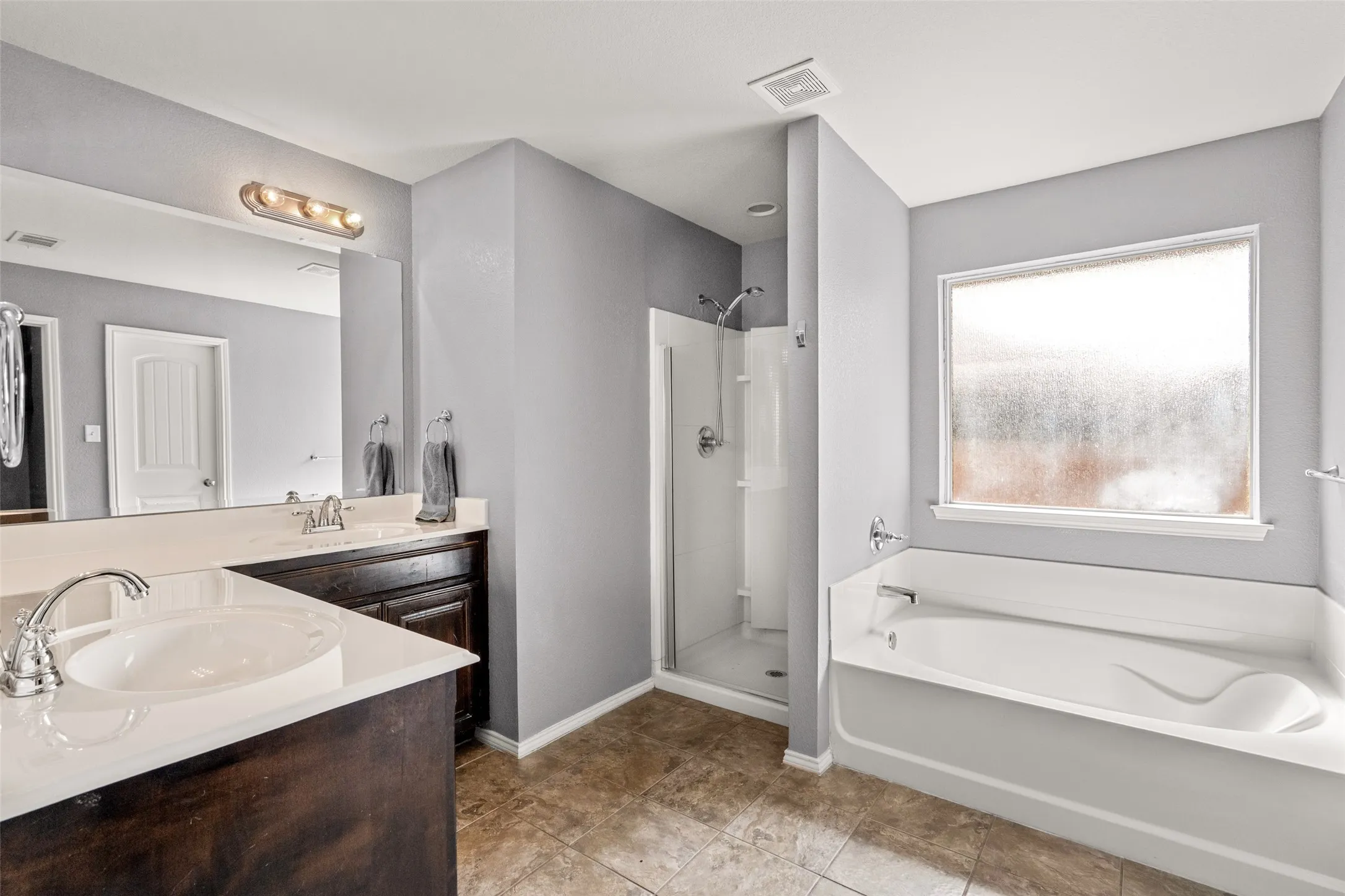Dual vanities with white countertops, a built-in shower with a glass enclosure, a white soaking tub, and tile flooring