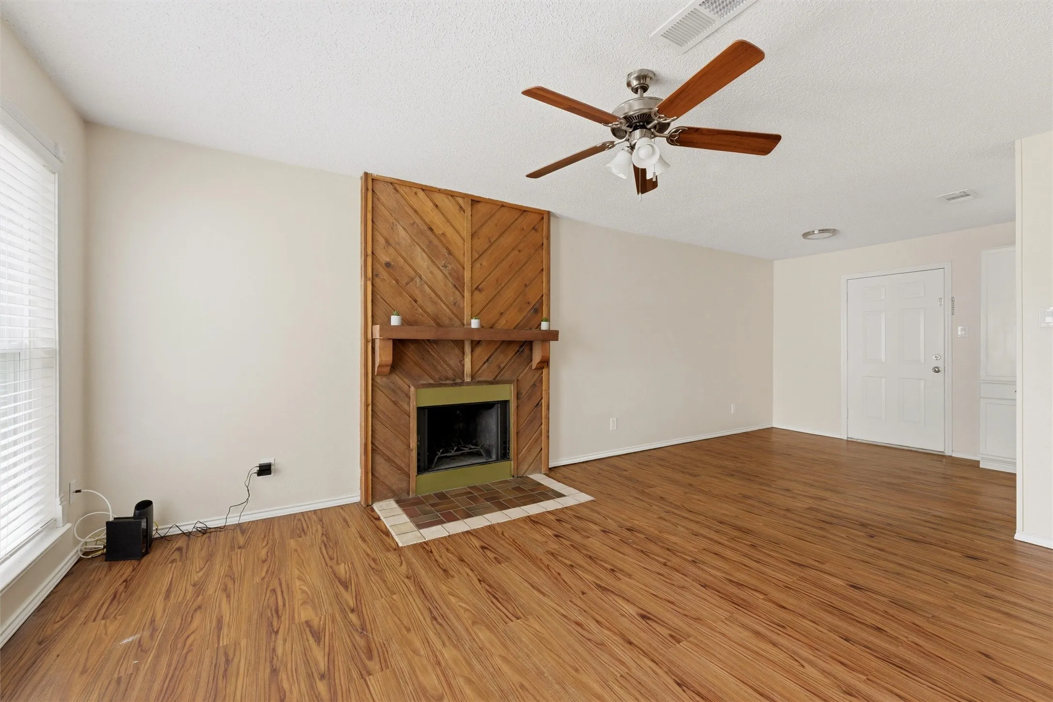 Wood-finish flooring, wood-paneled fireplace, built-in fireplace mantel, ceiling fan with integrated lighting, and a white entrance door
