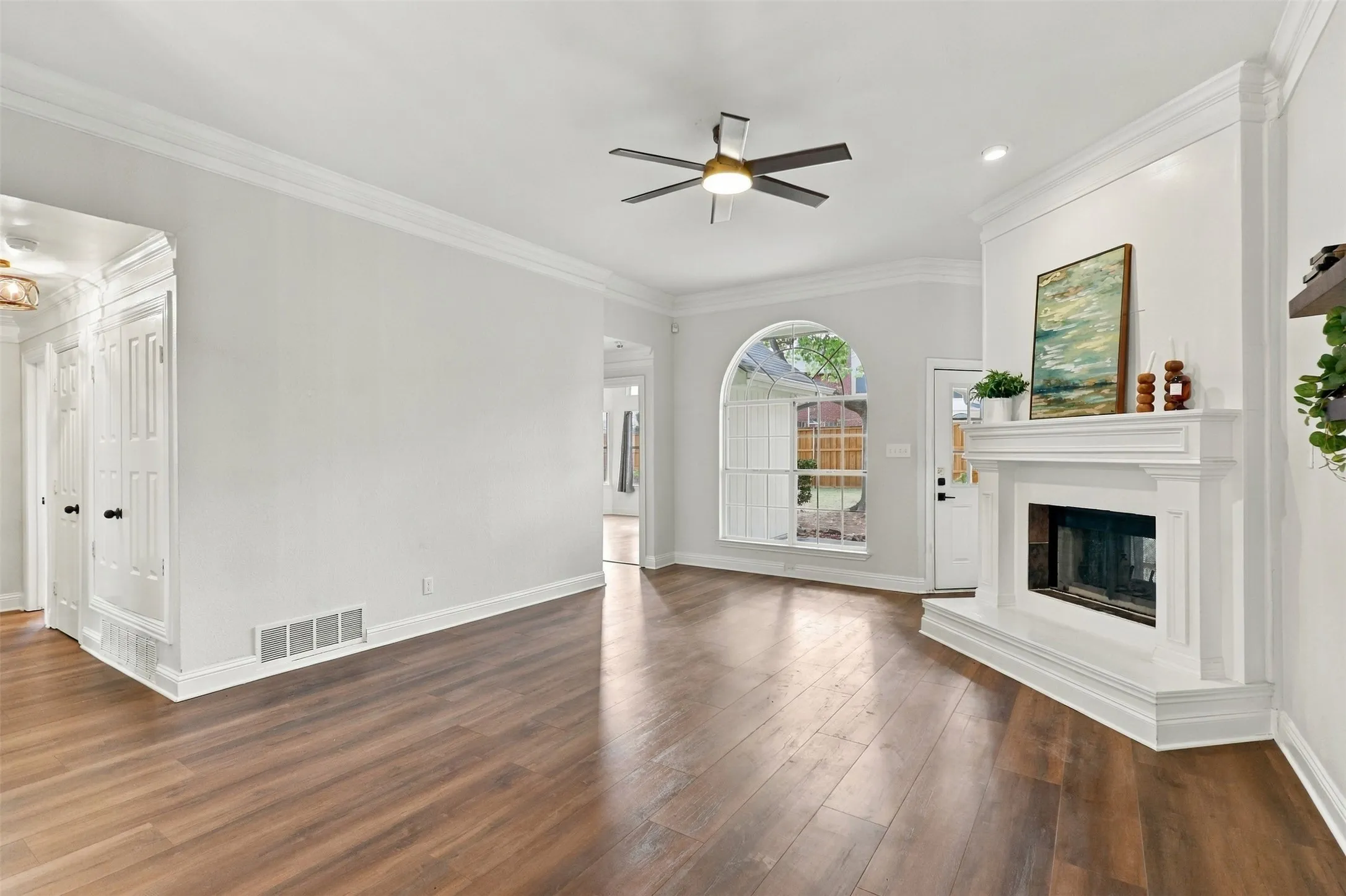Spacious living area featuring wood-finish flooring, a white mantel fireplace, an arched window, crown molding, and a ceiling fan
