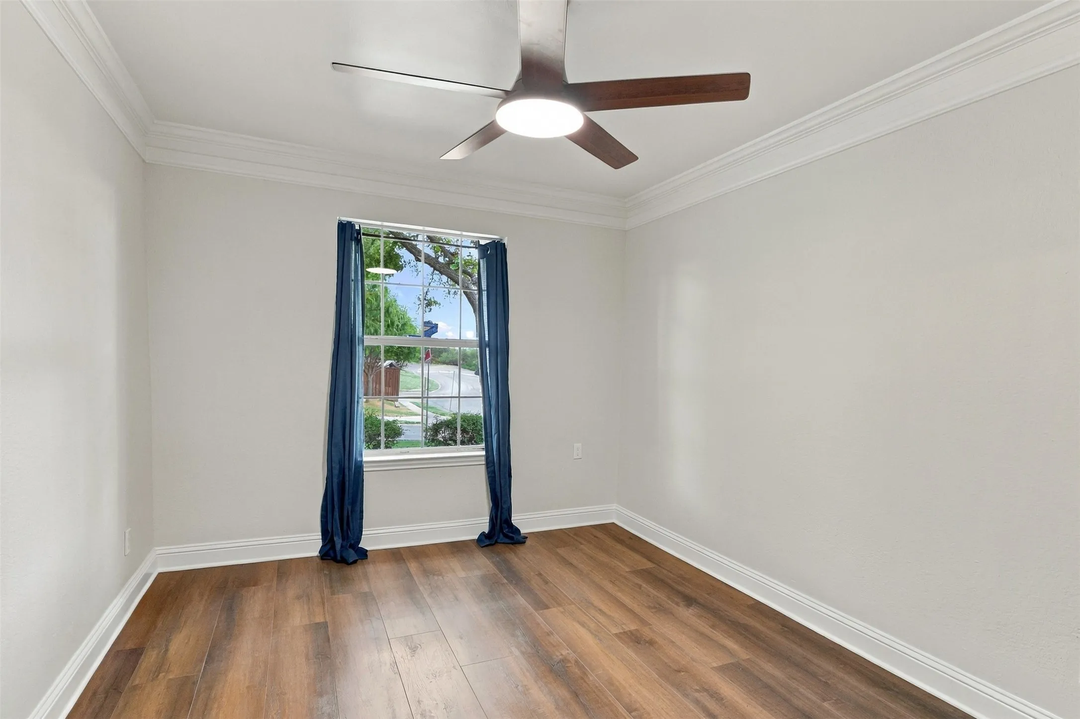 Wood-finish flooring, white painted walls, crown molding, a window with white trim, and a ceiling fan with integrated lighting