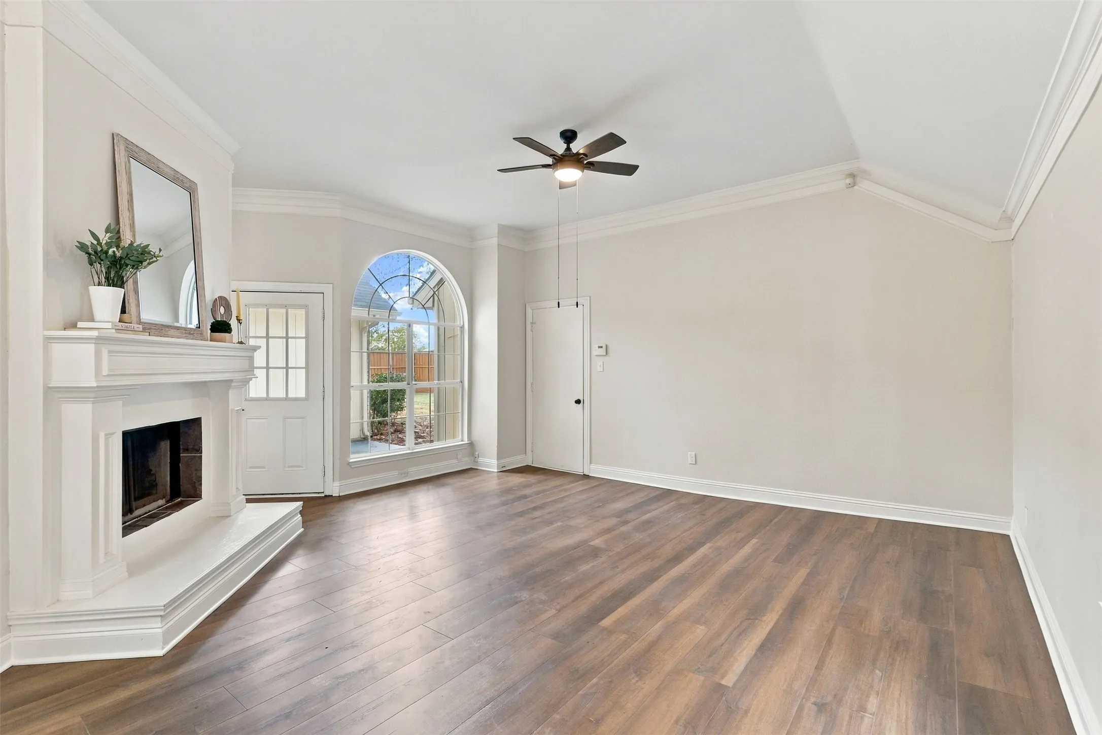 Wood-finish flooring throughout, white fireplace with hearth, arched window, vaulted ceiling, and a ceiling fan with integrated lighting