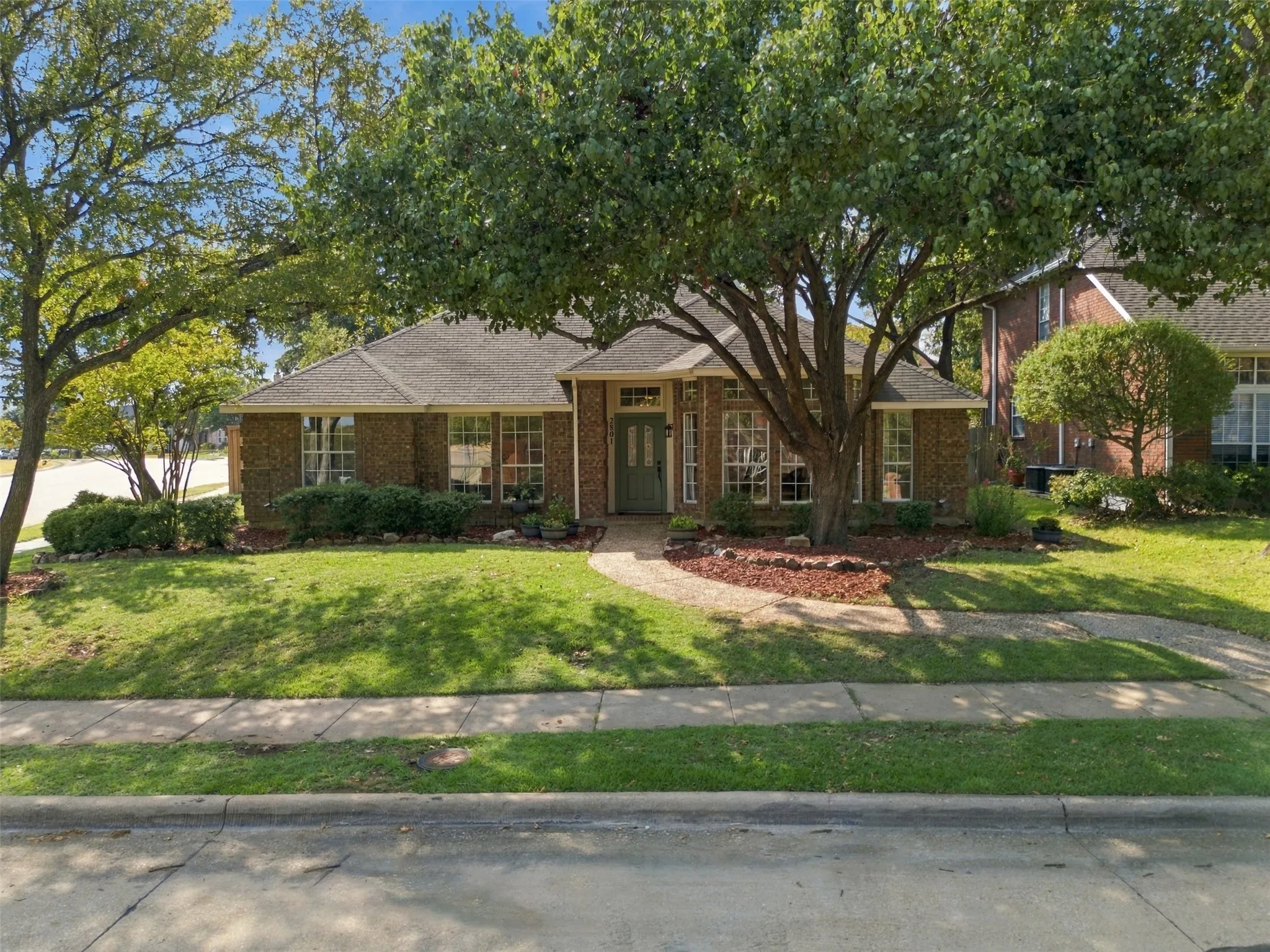Brick exterior with a gabled roof, mature shade trees, a concrete pathway, and landscaped shrubbery