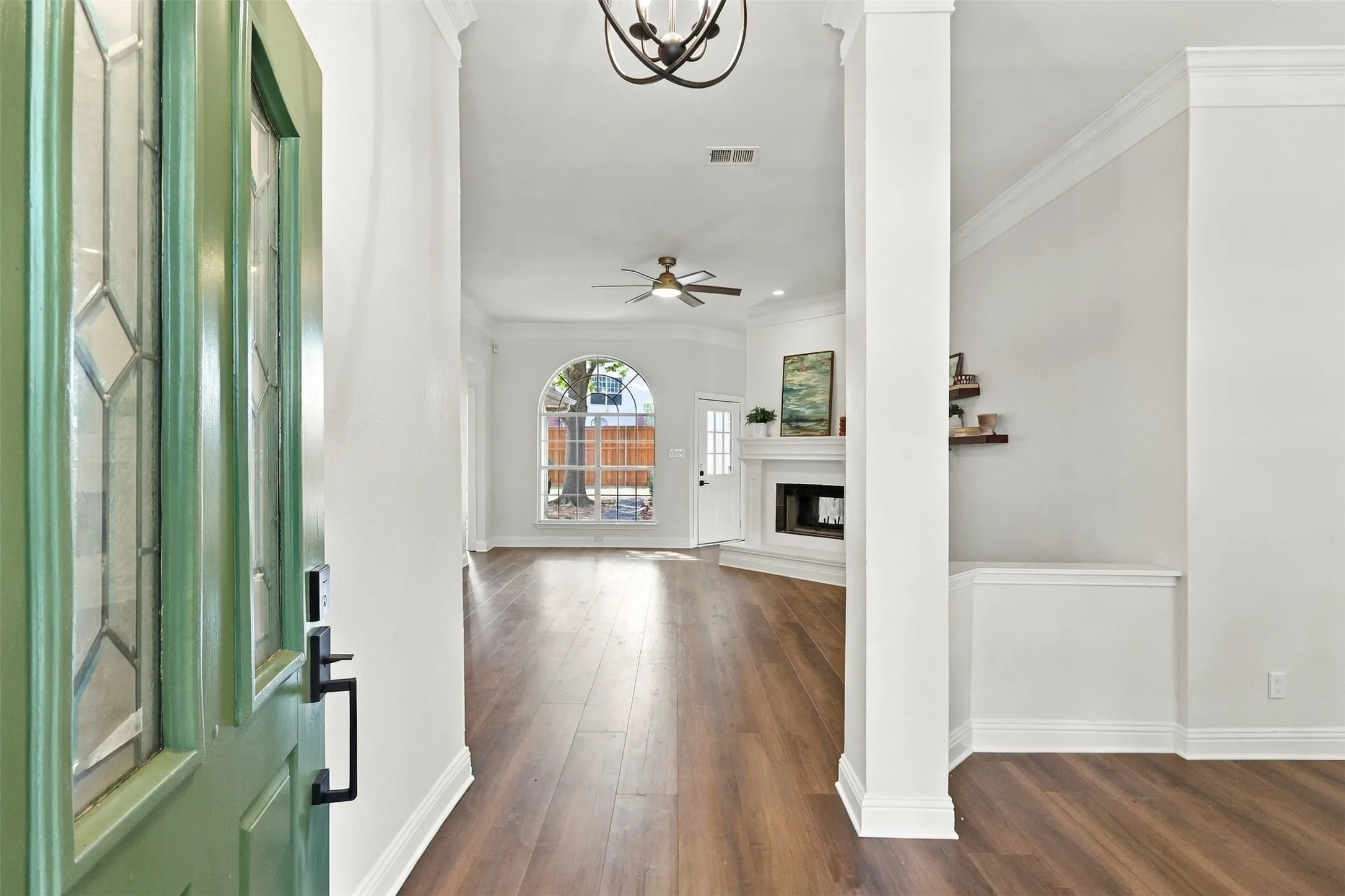 Green front door with leaded glass inserts, wide plank wood-finish flooring, white walls, arched window, and a built-in fireplace with mantle