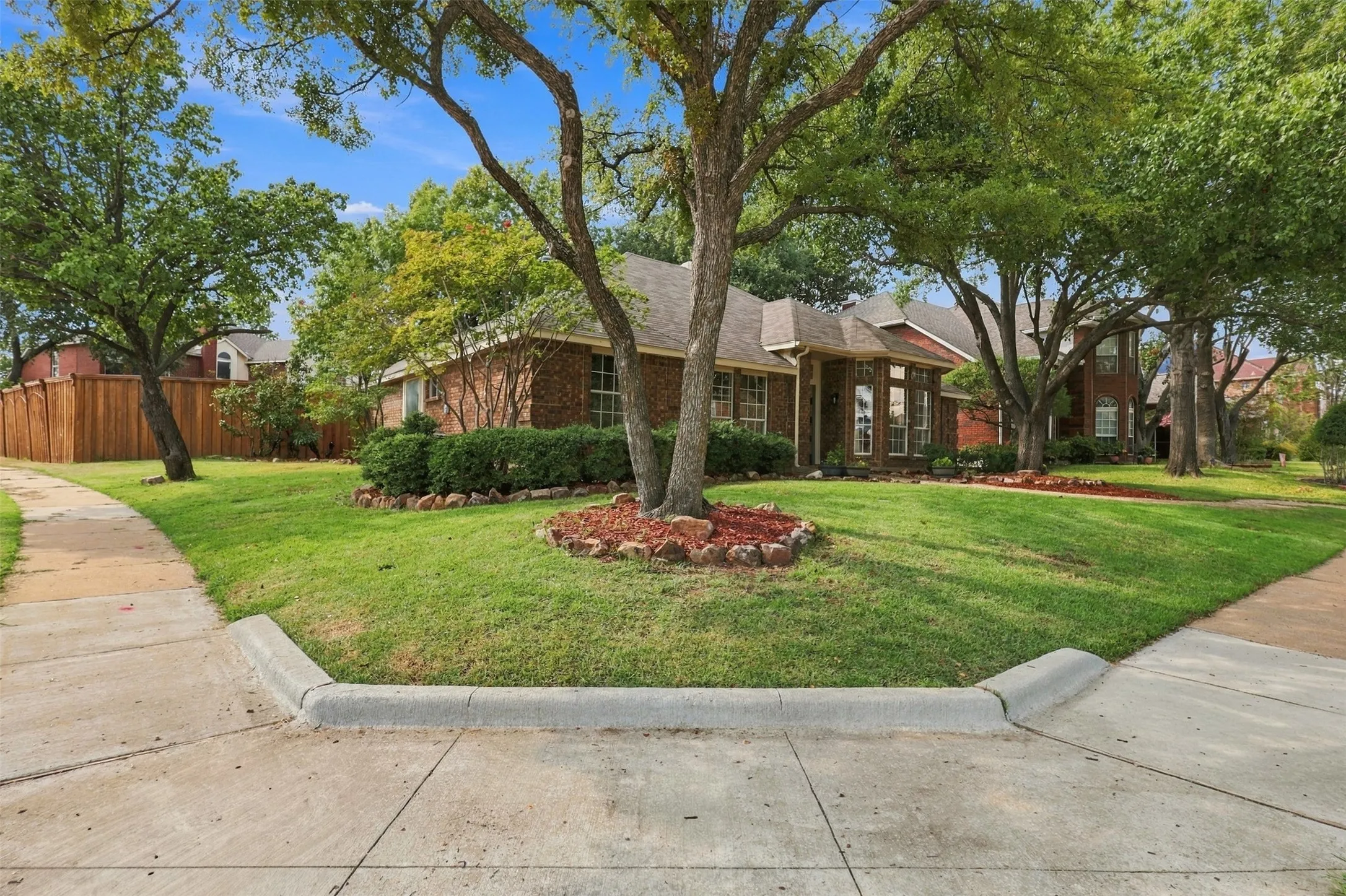 Brick exterior construction with multiple large windows, mature trees, landscaped beds, and a wood privacy fence