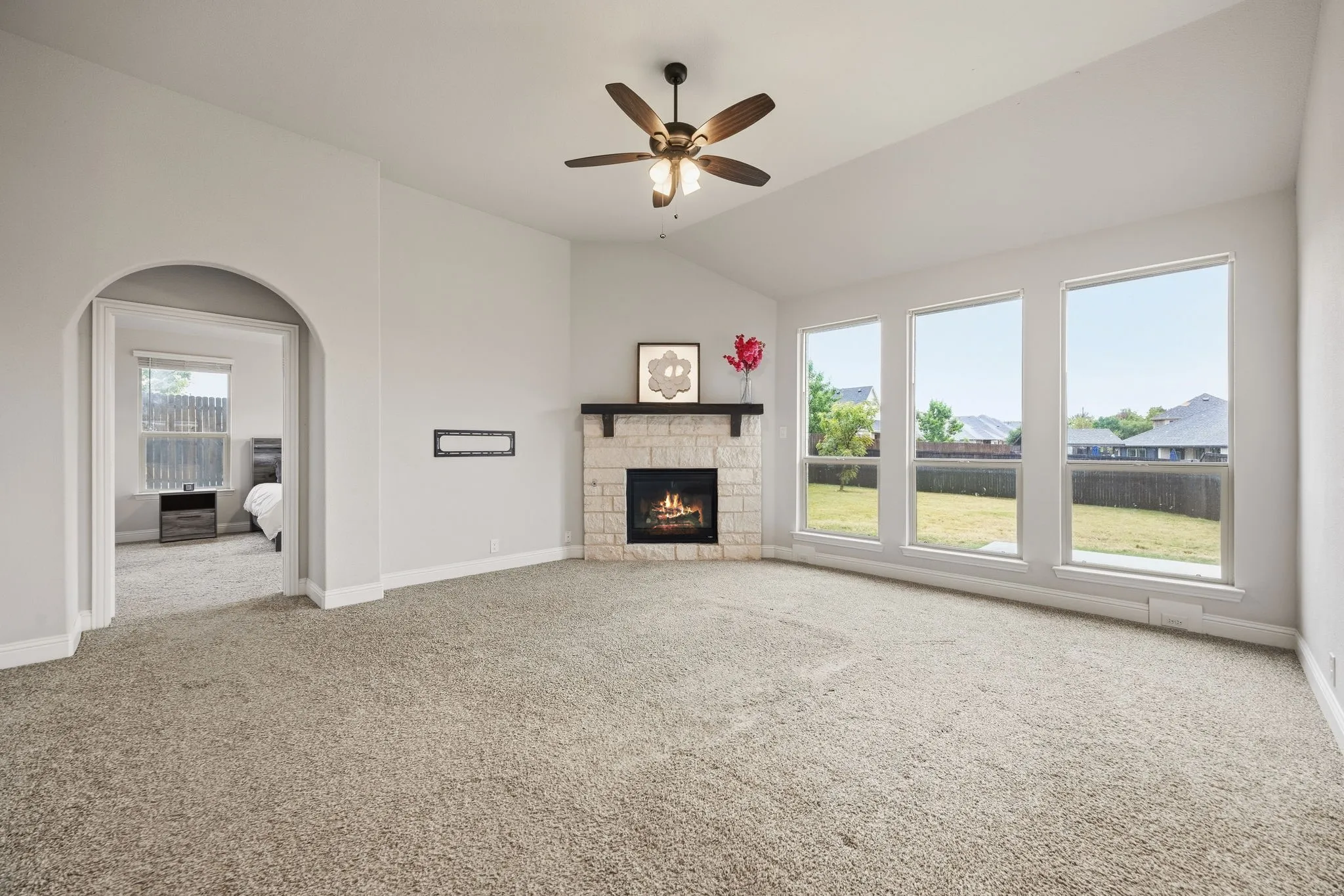 Vaulted ceiling, ceiling fan with integrated lighting, corner fireplace with stone surround and wood mantel, triple picture windows, and arched doorway