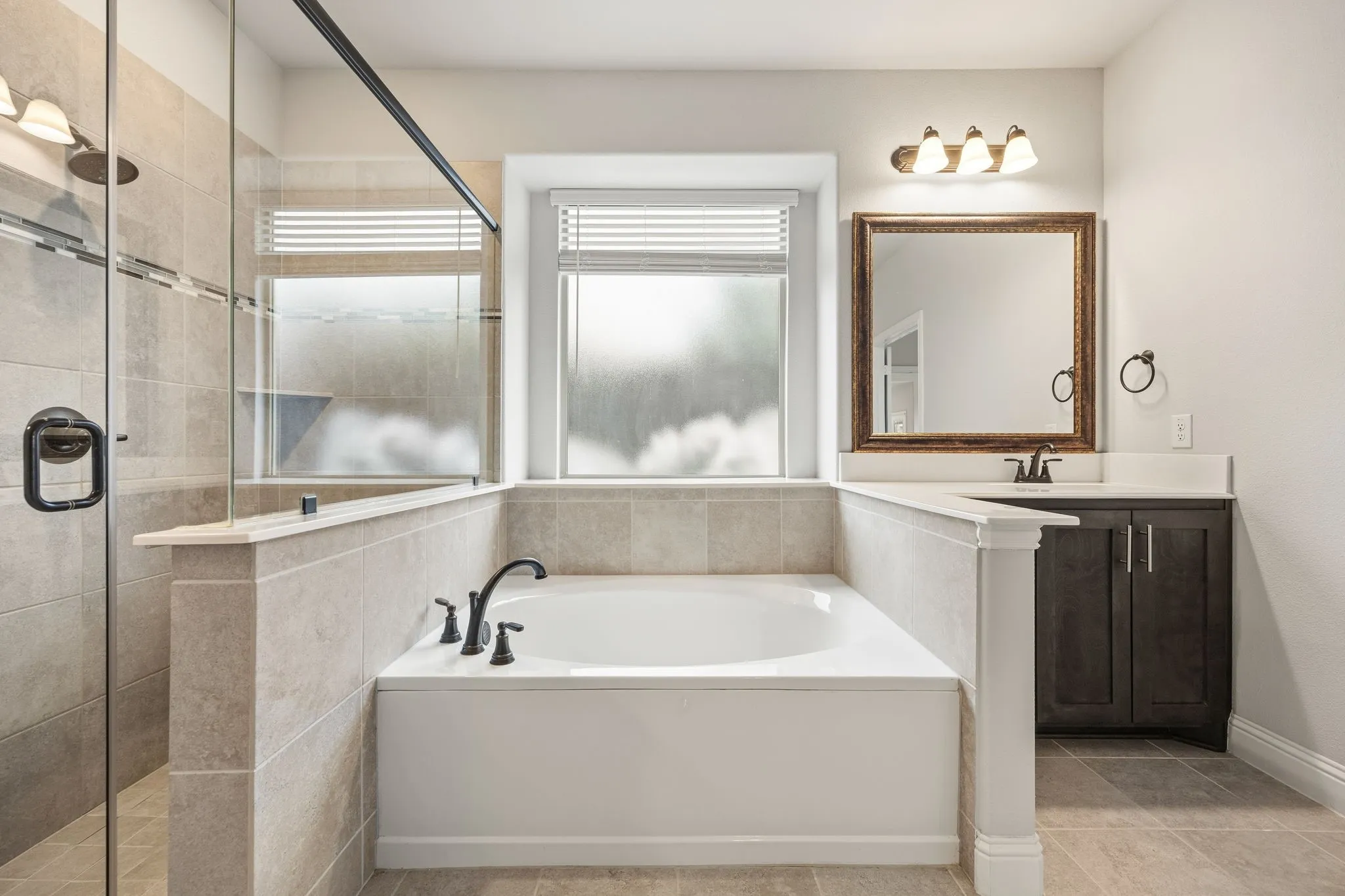 Built-in bathtub with black fixtures, glass-enclosed shower, tiled walls, single vanity with dark wood-finish cabinetry, and a large framed mirror