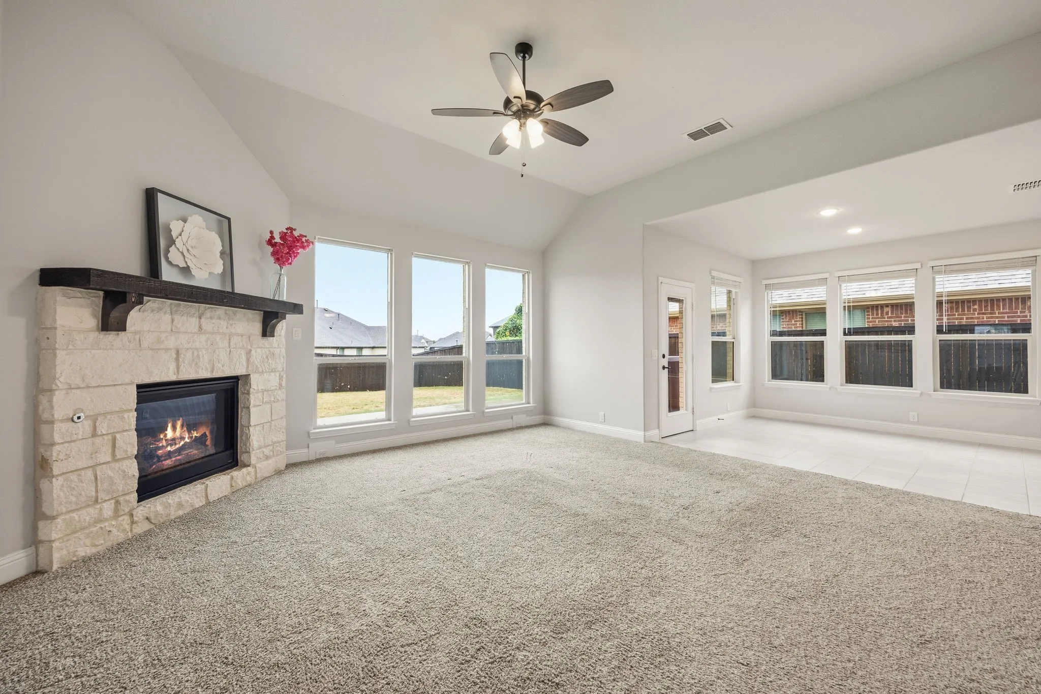 Vaulted ceilings with a ceiling fan, stone-clad fireplace with wood mantle, large windows with exterior views, light-toned carpet flooring, and tile flooring