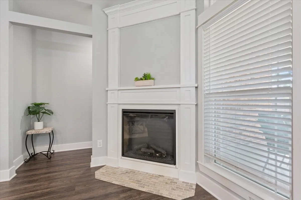 Corner fireplace with a white mantel and tile hearth, accompanied by wood-finish flooring and a window with white blinds