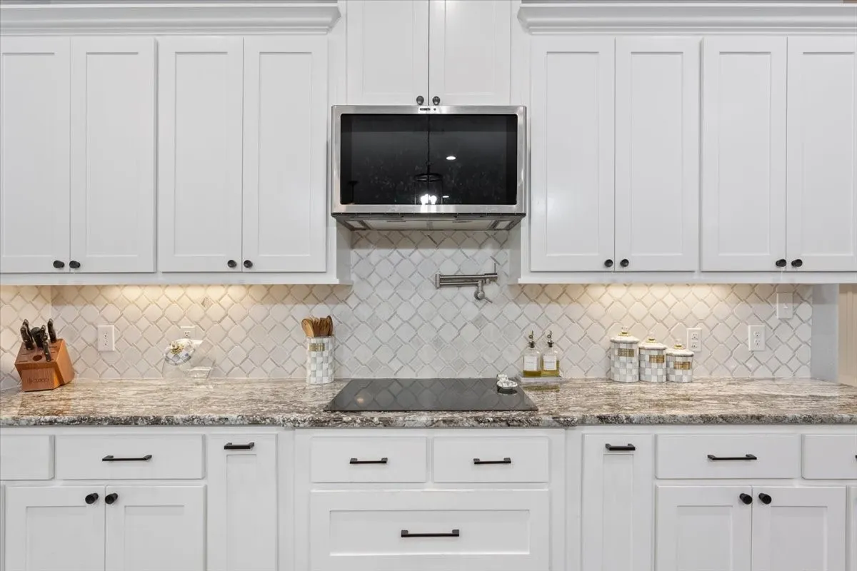 White shaker-style cabinetry, patterned tile backsplash, built-in stainless steel microwave, stone countertops, and black electric cooktop