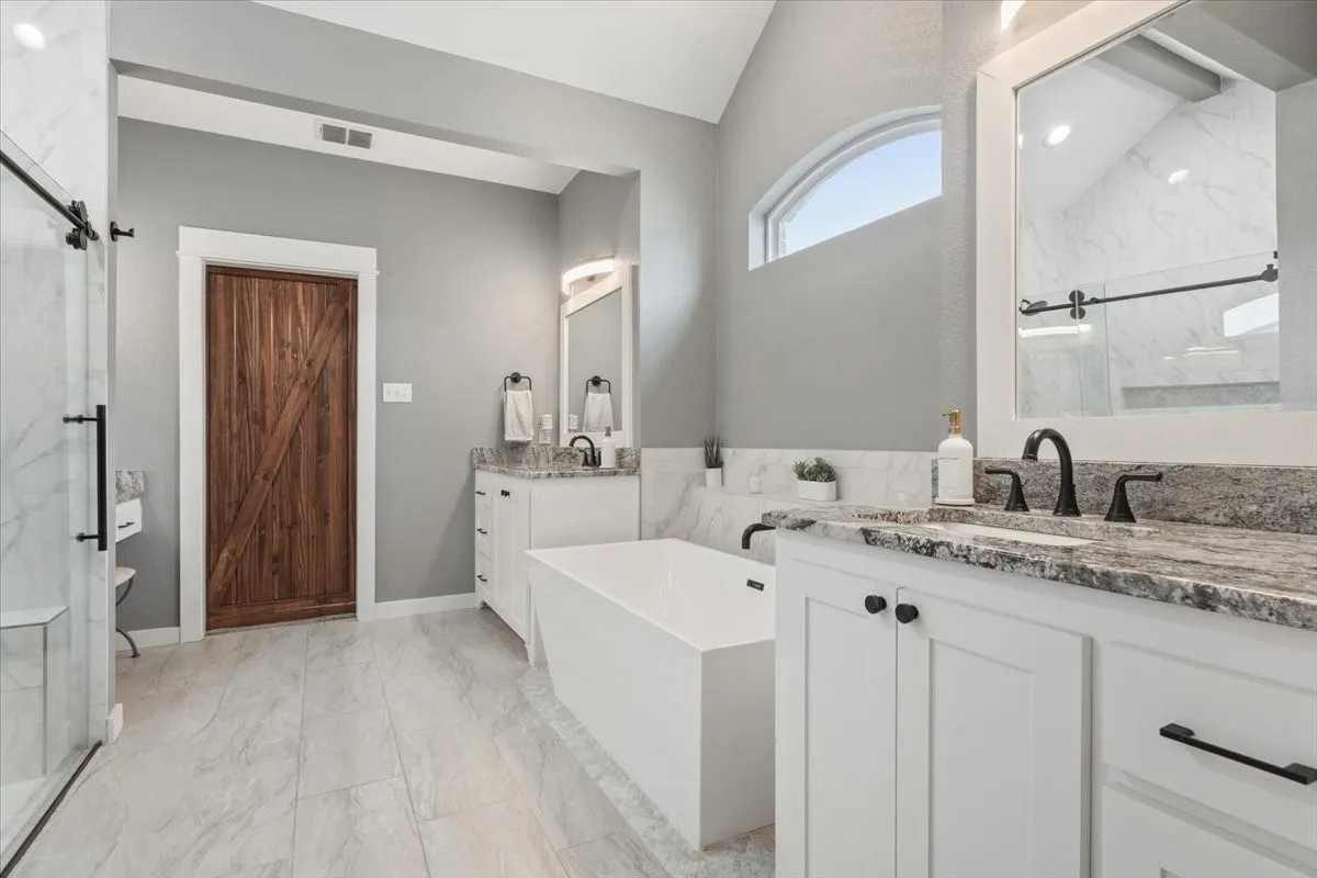 Vaulted ceiling, freestanding soaking tub, dual vanities with stone countertops, arched transom window, and a sliding glass shower enclosure