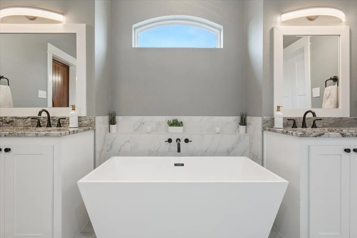 Freestanding soaking tub with wall-mounted faucet, dual vanities with granite countertops, and an arched transom window