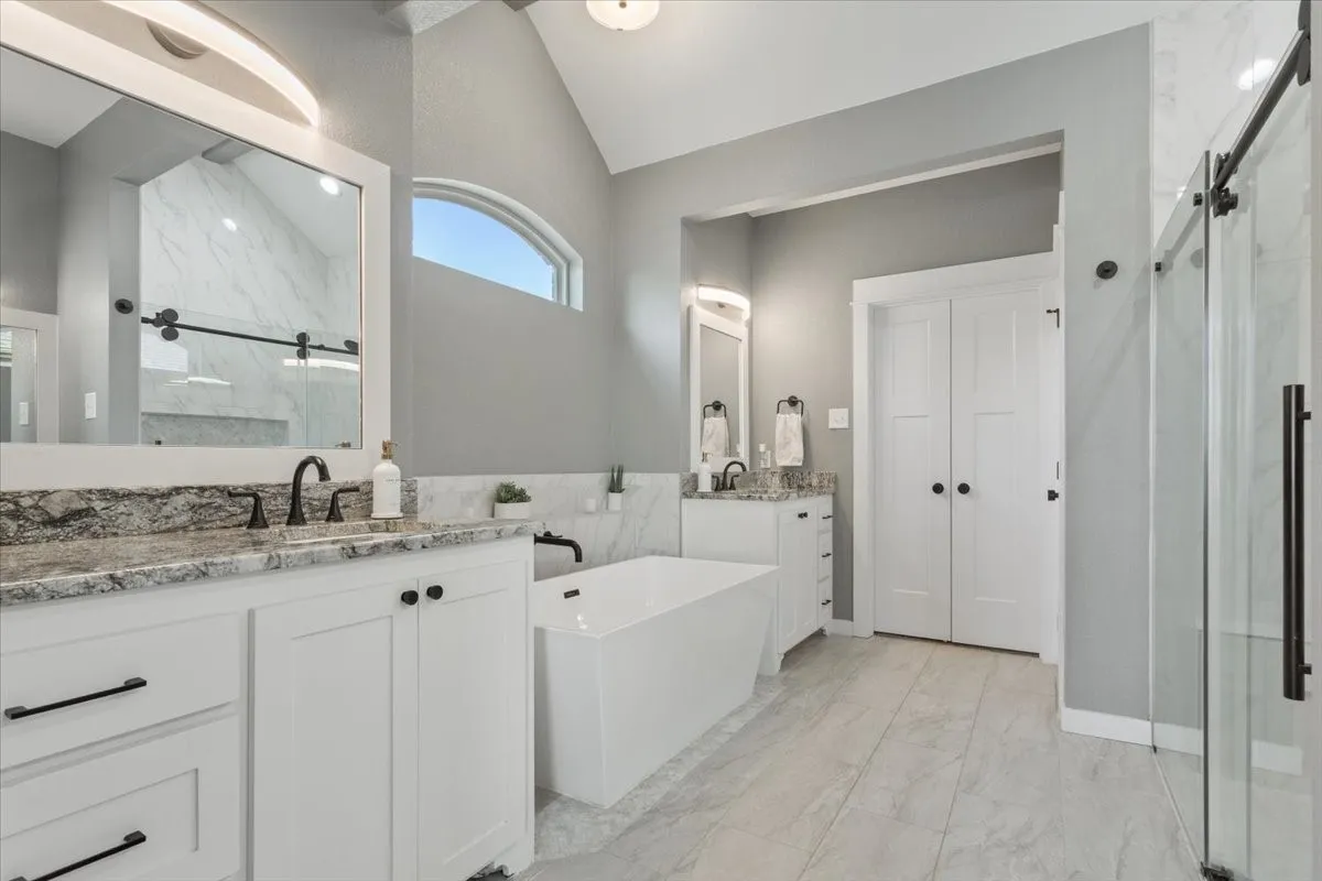 Dual vanities with stone countertops, a freestanding soaking tub, a walk-in shower with a sliding glass door, an arched transom window, and vaulted ceilings