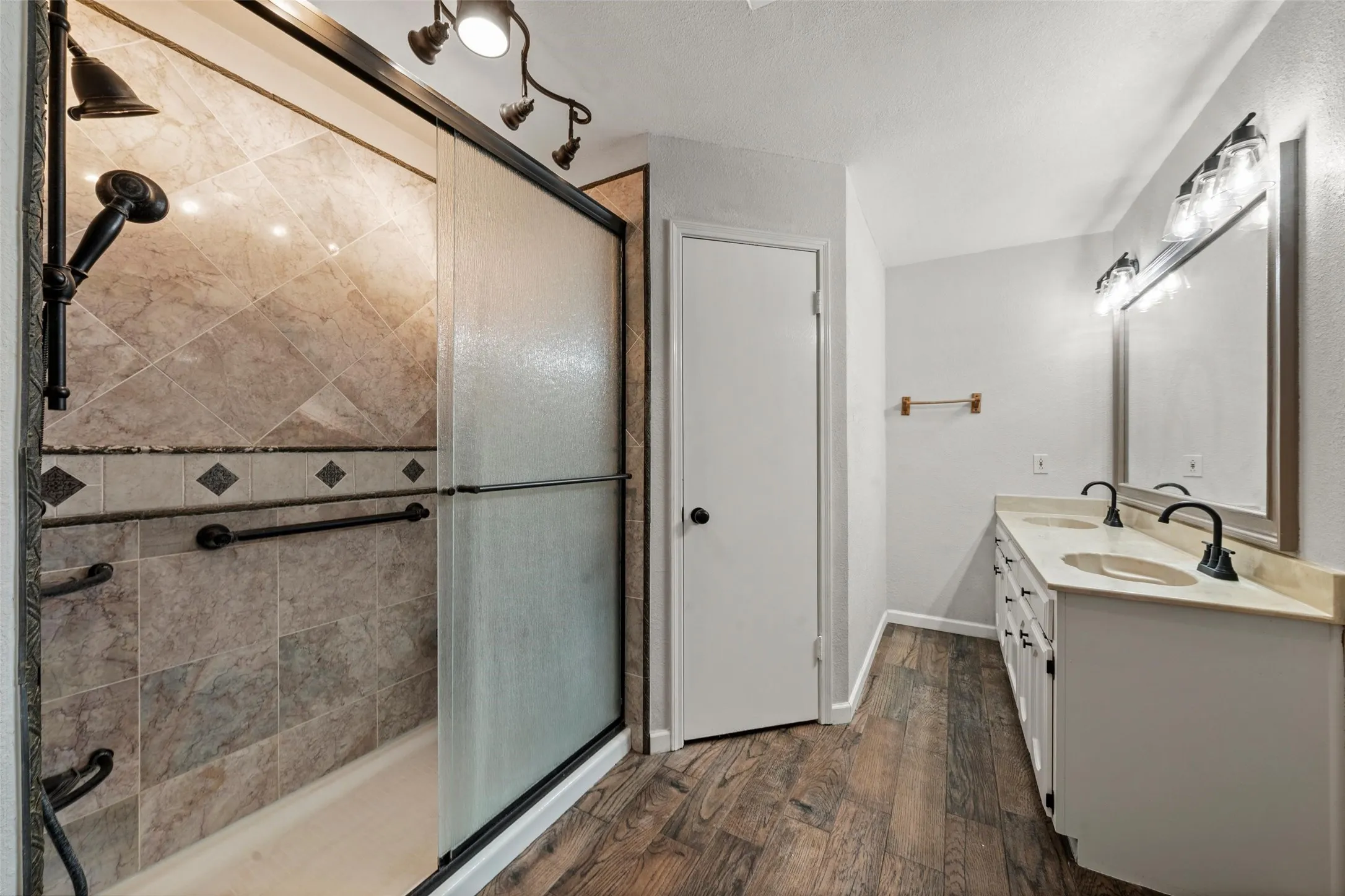 Walk-in shower with tile surround and dark bronze fixtures, dual vanity with undermount sinks and light-toned countertops, wood-finish flooring, and track lighting