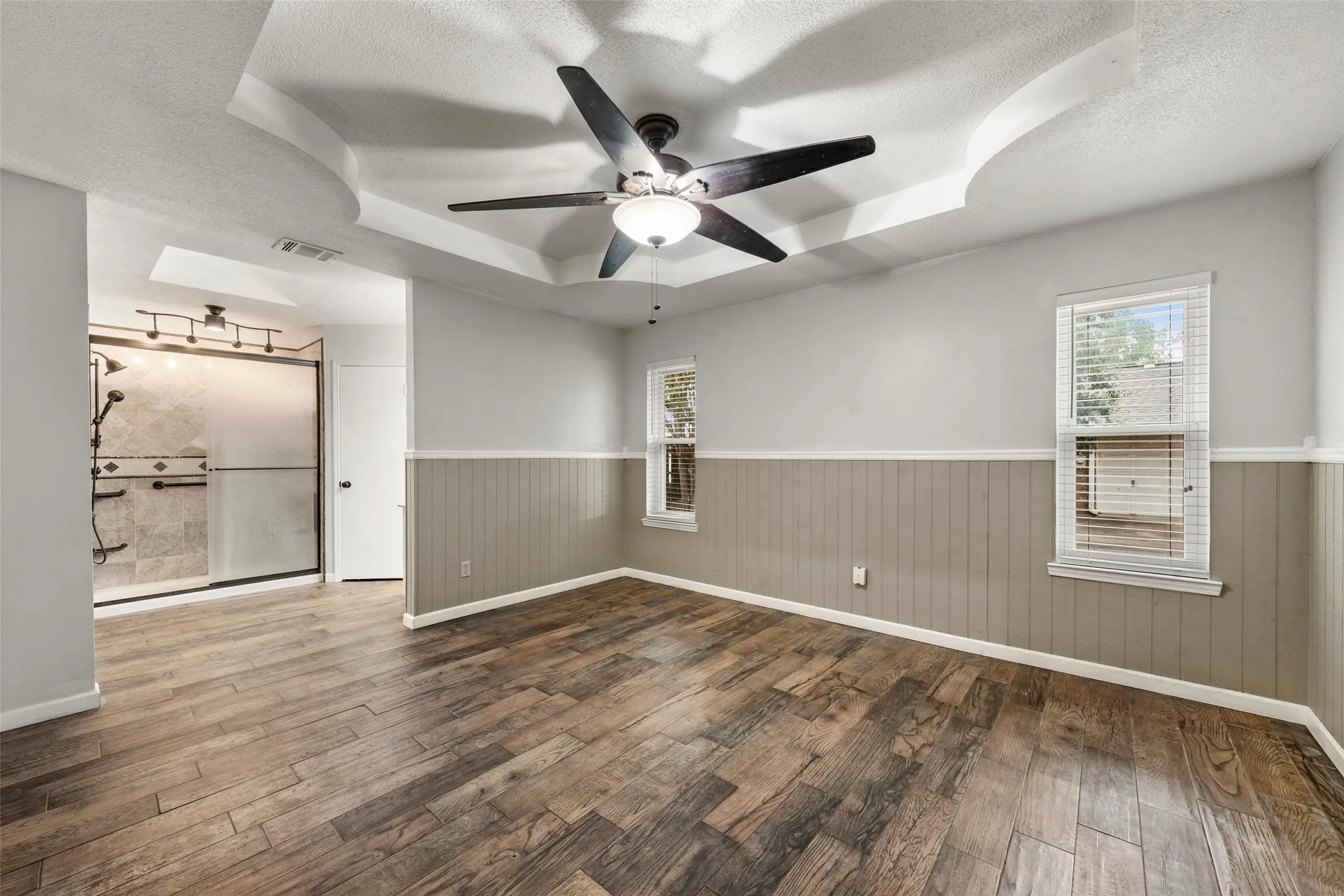 Tray ceiling with a ceiling fan, wood-finish flooring, two windows with blinds, wainscoting, and an adjacent shower with a glass enclosure