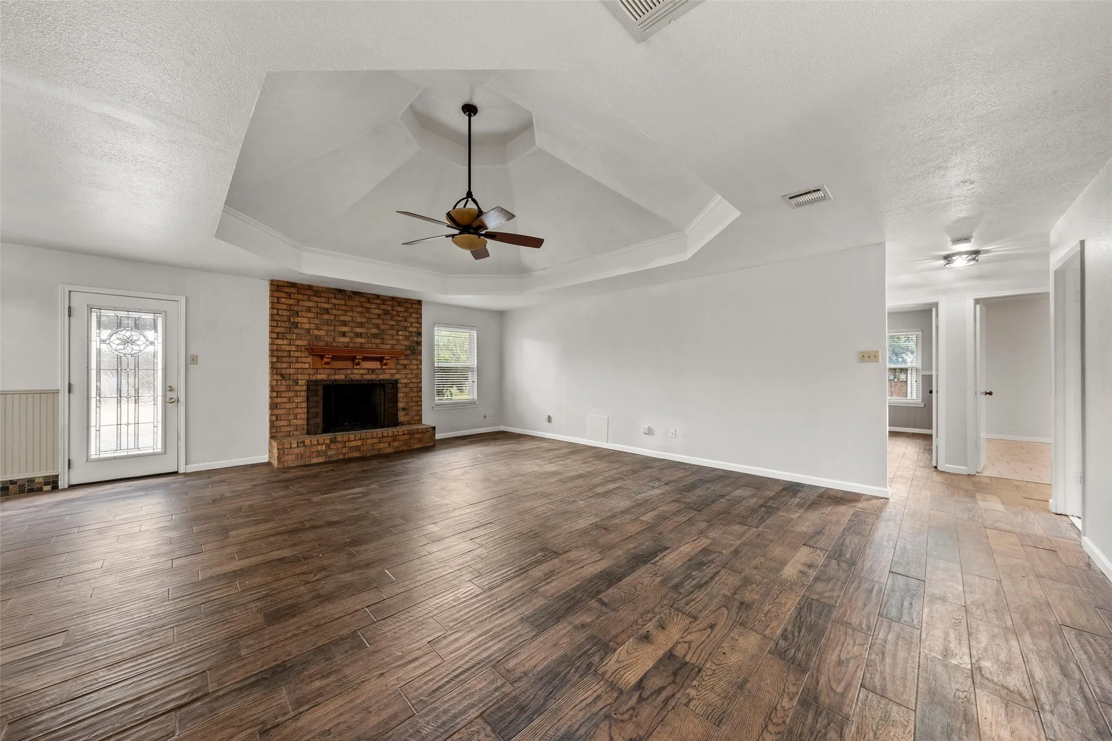 Tray ceiling with a ceiling fan, brick-faced fireplace with a wood mantel, wood-finish flooring, and white walls