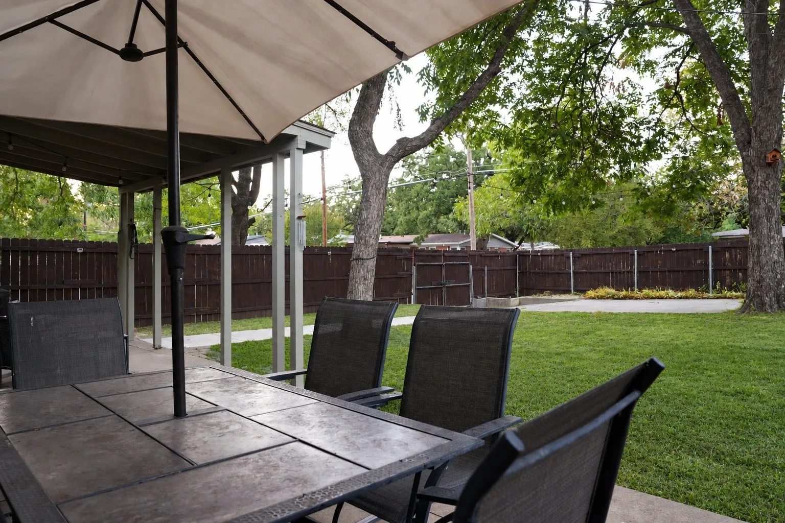 Covered patio area with a tiled surface, expansive lawn, mature trees, and a privacy fence