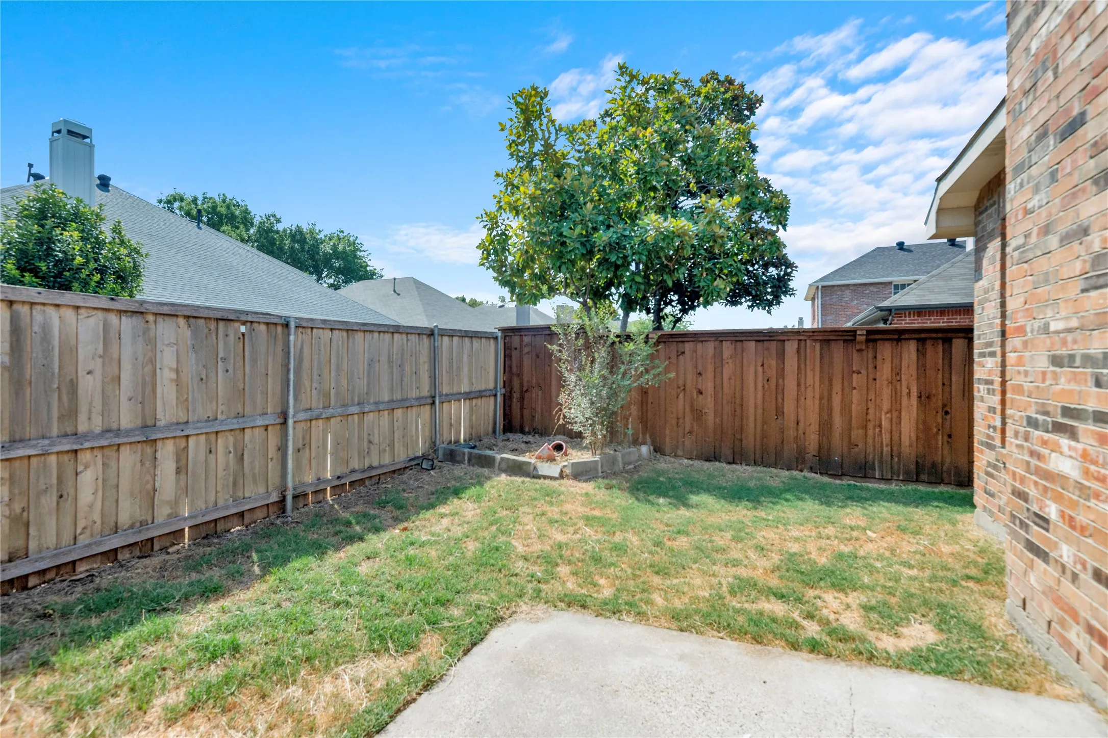 Landscaped rear yard featuring a mature tree, wood fencing, and a concrete patio