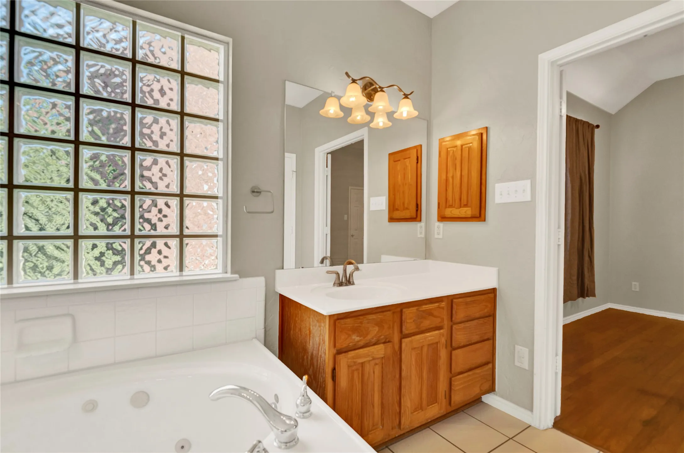 Deep soaking tub with chrome fixtures, glass block window, wood vanity with integrated sink, and wall-mounted light fixture
