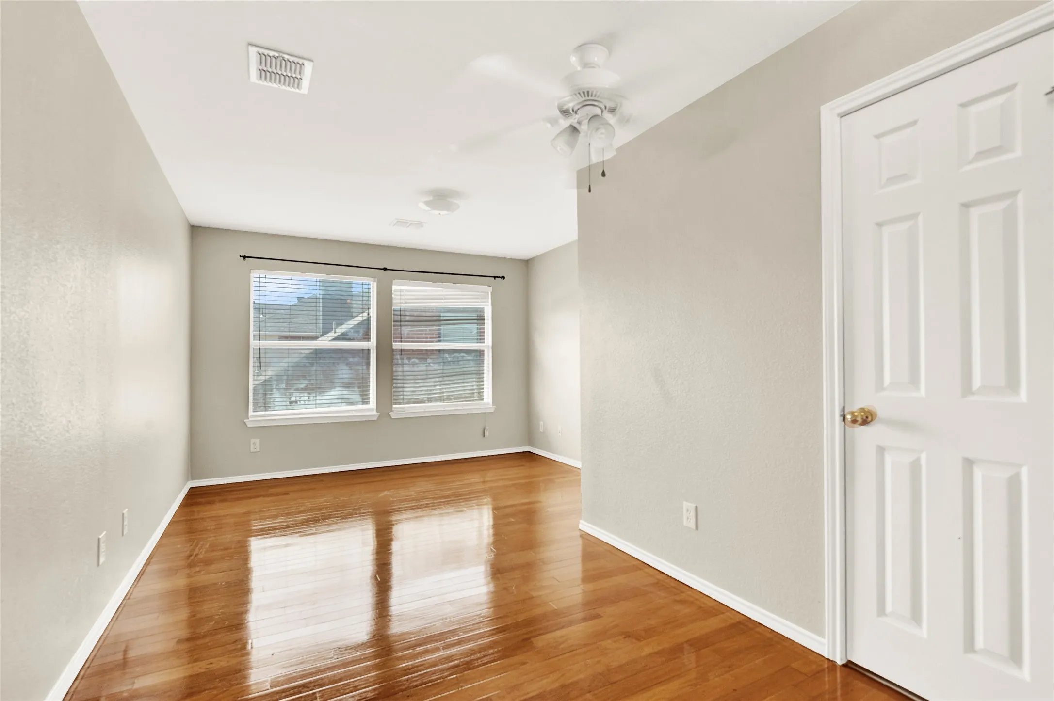 Wood-finish flooring with a high-gloss finish, two windows with blinds, a ceiling fan, and a paneled white door