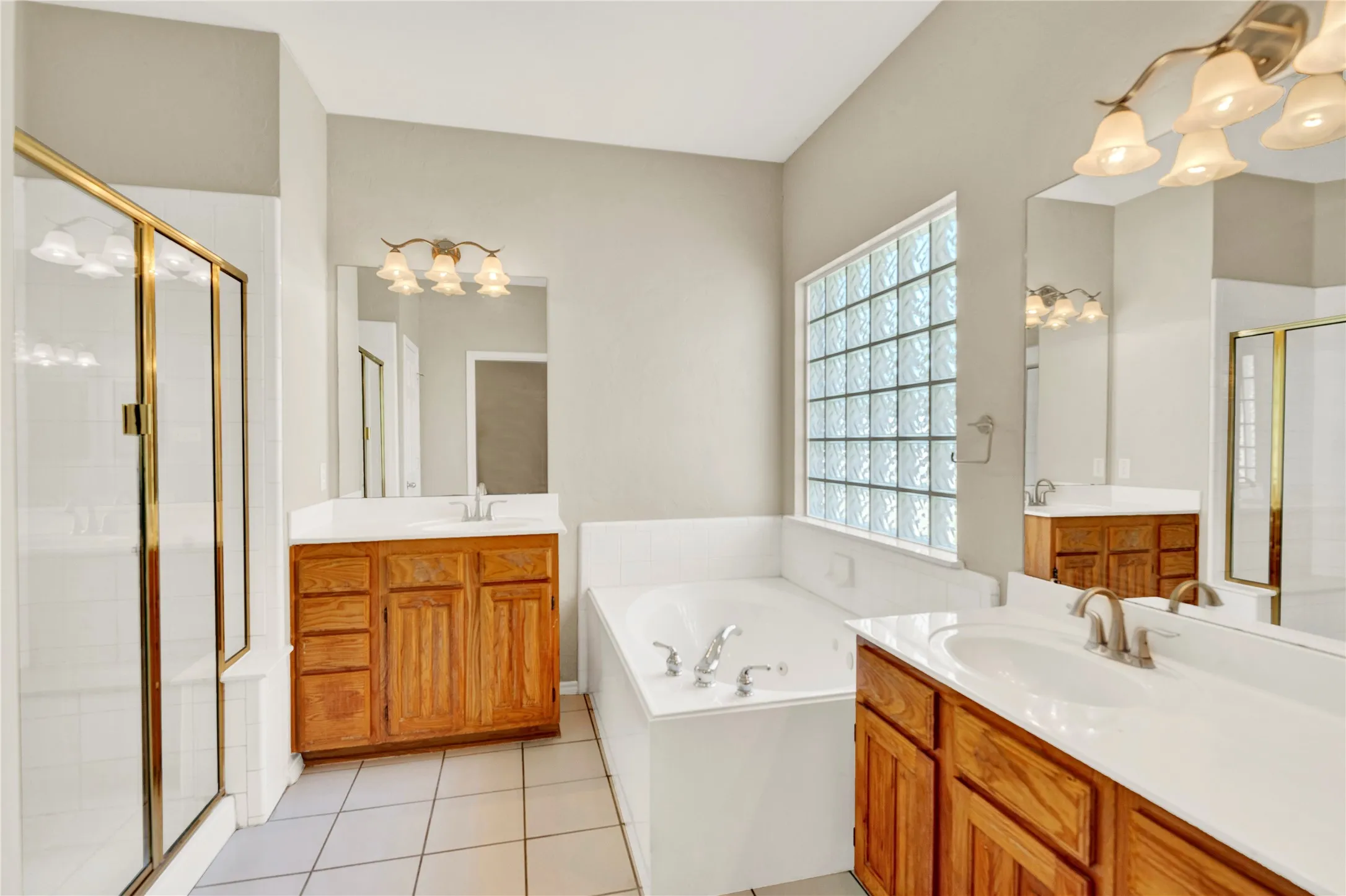 Dual wood-finish vanities with white countertops, corner soaking tub, glass block window, and a separate shower with a brass-finish frame