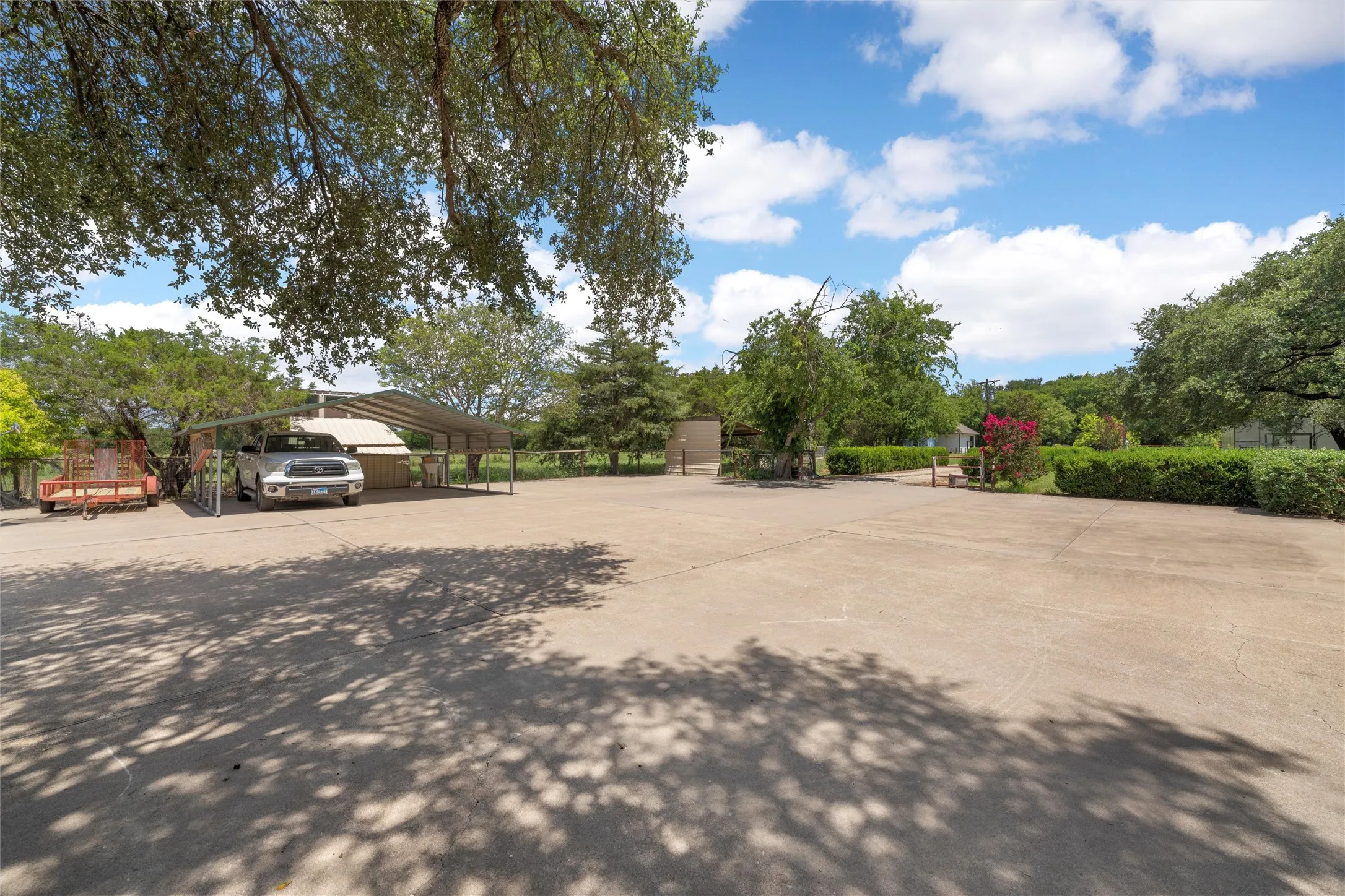 Expansive concrete parking area with a multi-vehicle carport and surrounding mature trees providing natural shade