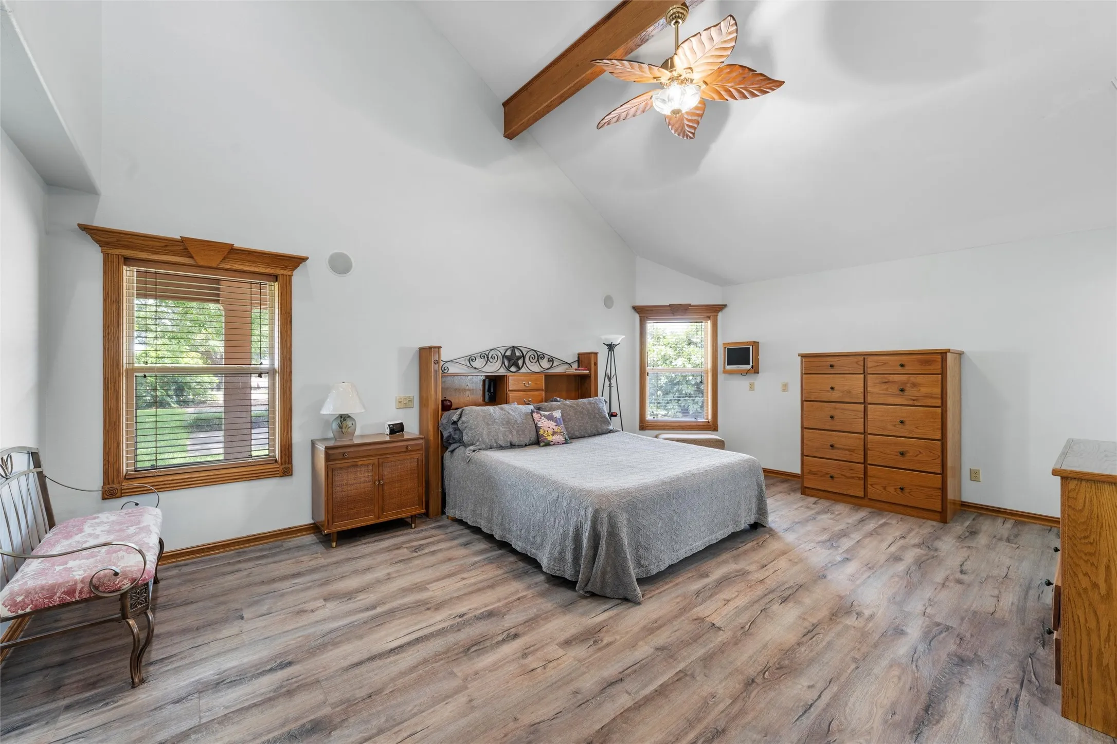 Spacious primary bedroom suite featuring vaulted ceilings with exposed wooden beams, wood-finish flooring throughout, and abundant natural light from multiple windows