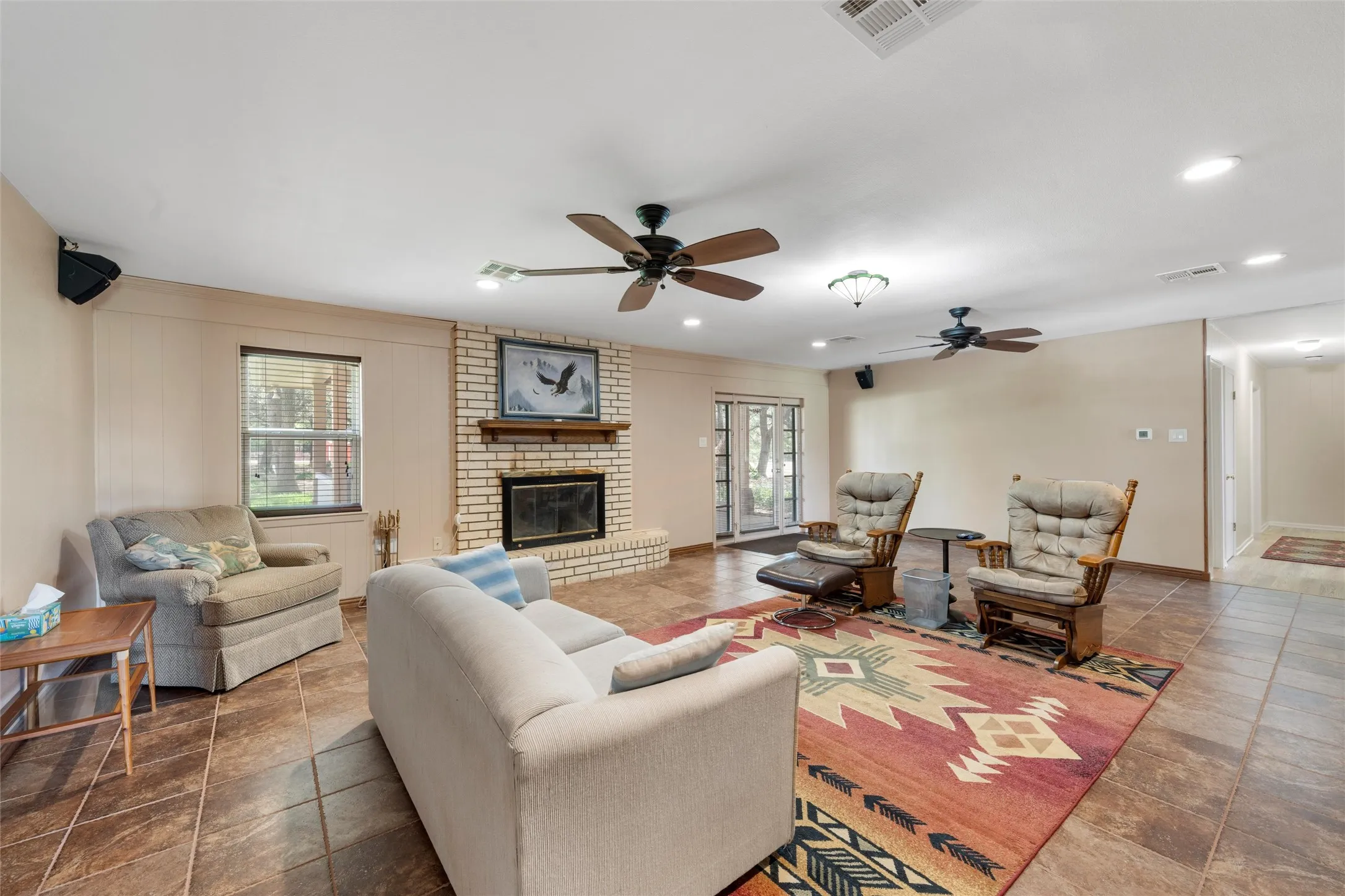 Spacious room featuring tile flooring, a brick fireplace with a wood mantle, and multiple ceiling fans