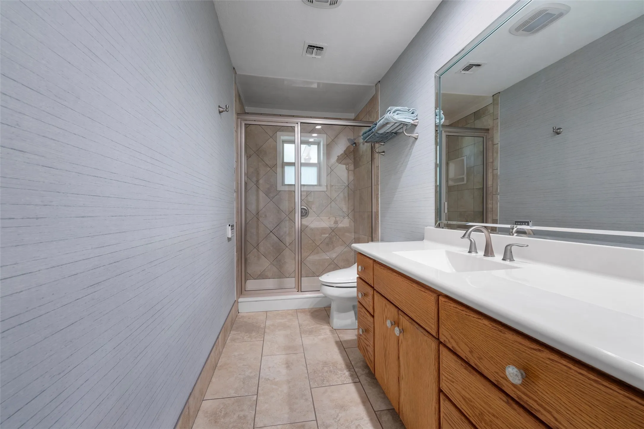 Double vanity with integrated sinks and brushed nickel faucets, wood-finish cabinetry, frameless mirror, tiled flooring, and a glass-enclosed shower with tiled surround