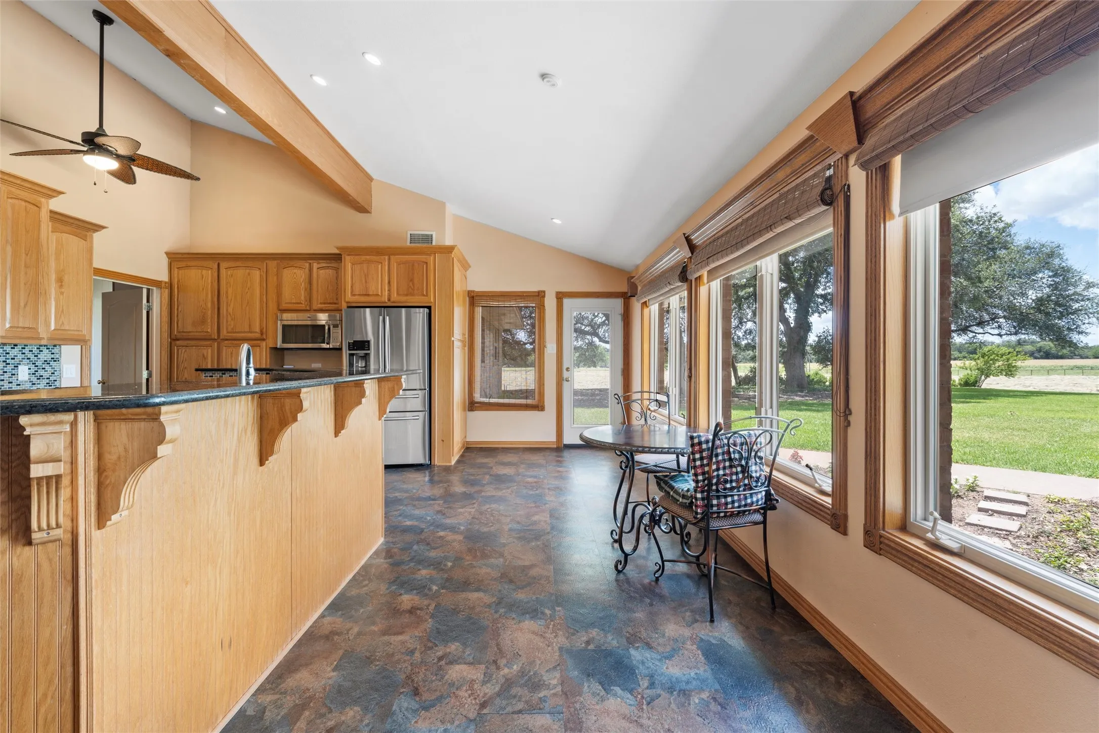 Vaulted ceiling with exposed wood beams, wood cabinetry, stainless steel appliances, large windows with roller shades, and a glass-paneled door