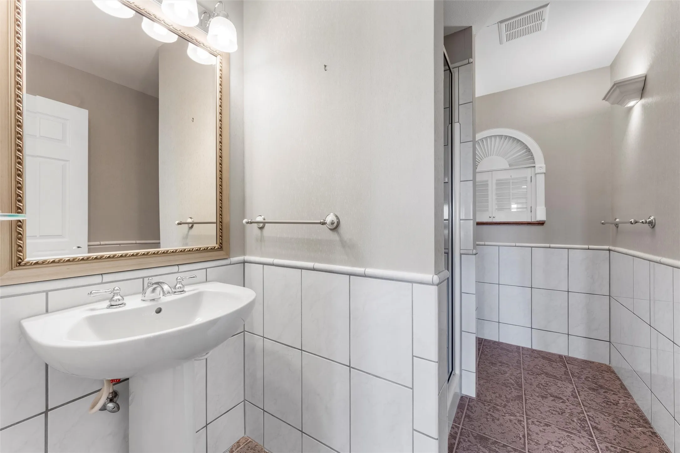 Pedestal sink with chrome fixtures, framed mirror, wall-mounted light fixture, and white tiled wainscoting