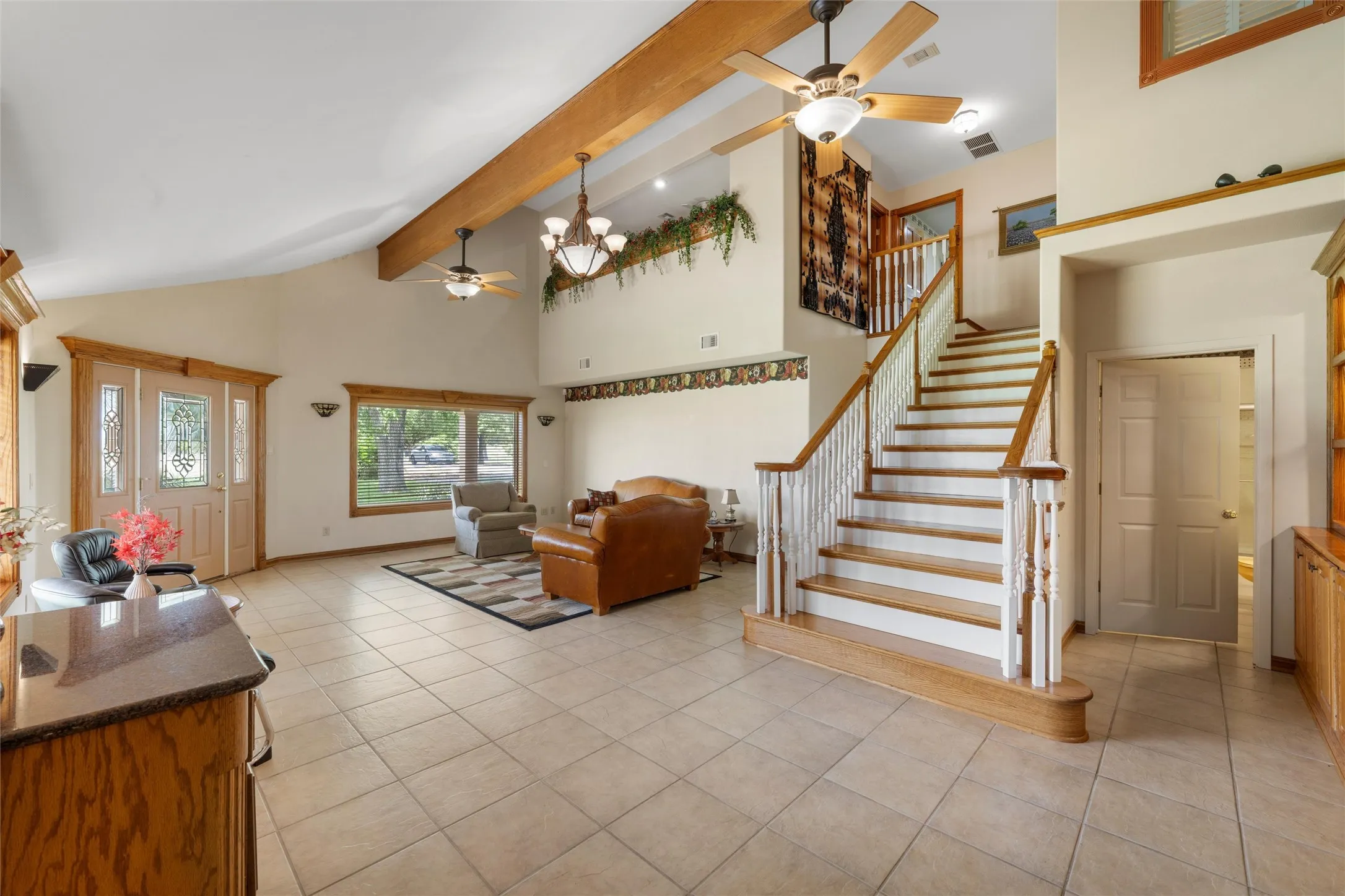 Vaulted ceilings with exposed wood beams, tile flooring, wood-finish staircase with white risers and balusters, and a decorative front entry door with sidelights