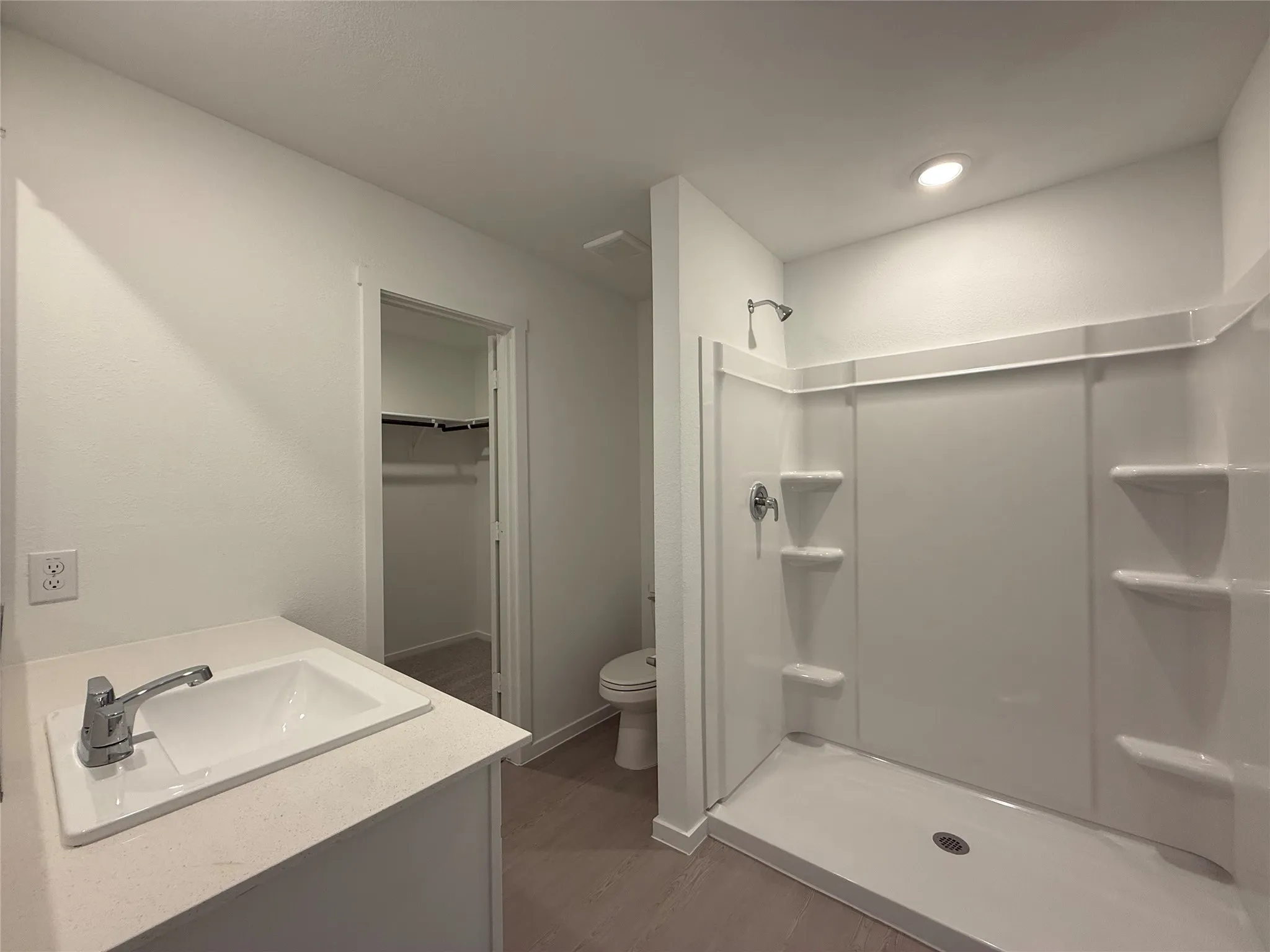White vanity with integrated sink and chrome faucet, white shower stall with built-in shelving, white toilet, and wood-finish flooring