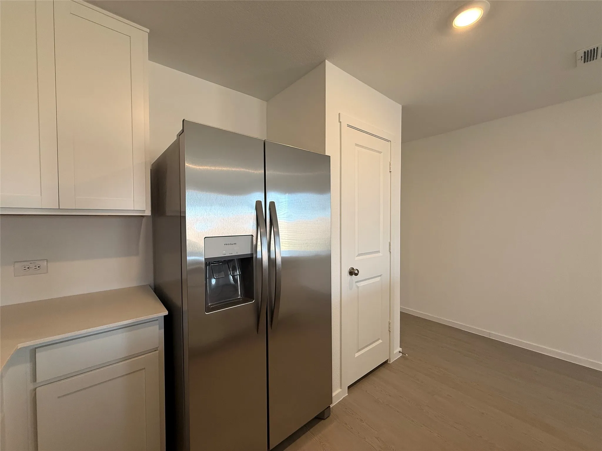 Stainless steel refrigerator, white shaker-style cabinetry, light-toned countertop, wood-finish flooring, and recessed ceiling lighting