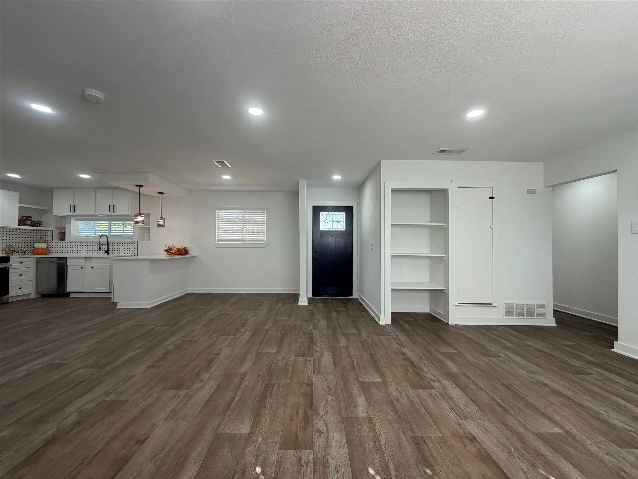 Open-concept living area featuring wood-finish flooring, recessed lighting, built-in shelving, a modern kitchen with white cabinetry, and a dark-paneled entry door with decorative glass