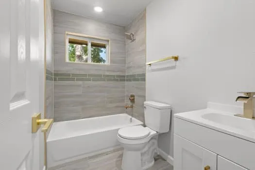 White paneled door with brass hardware, tub with tile surround and glass accent, recessed lighting, white vanity with integrated sink, and brass faucet