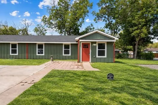 Green exterior siding with white trim, a red entry door, a paved driveway, a stone paver patio, and a manicured lawn
