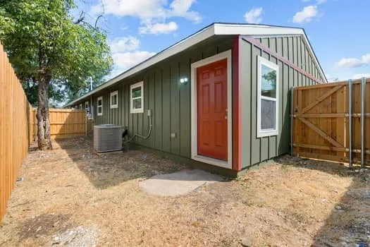 Green vertical siding exterior with a contrasting orange entry door, white trim windows, and a wood privacy fence