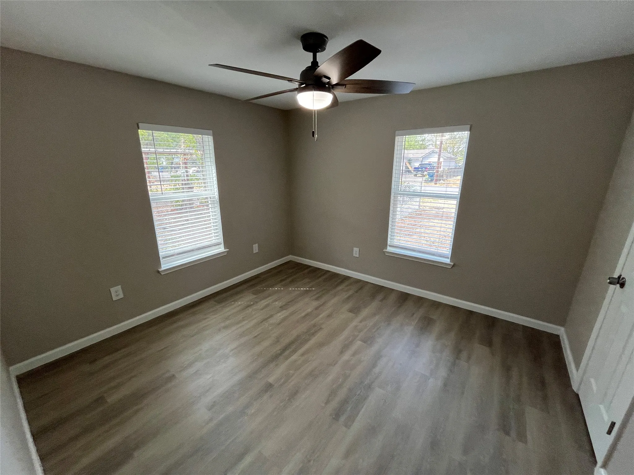 Neutral tone walls, wood-finish flooring, two windows with blinds, a ceiling fan with integrated lighting, and a white paneled door