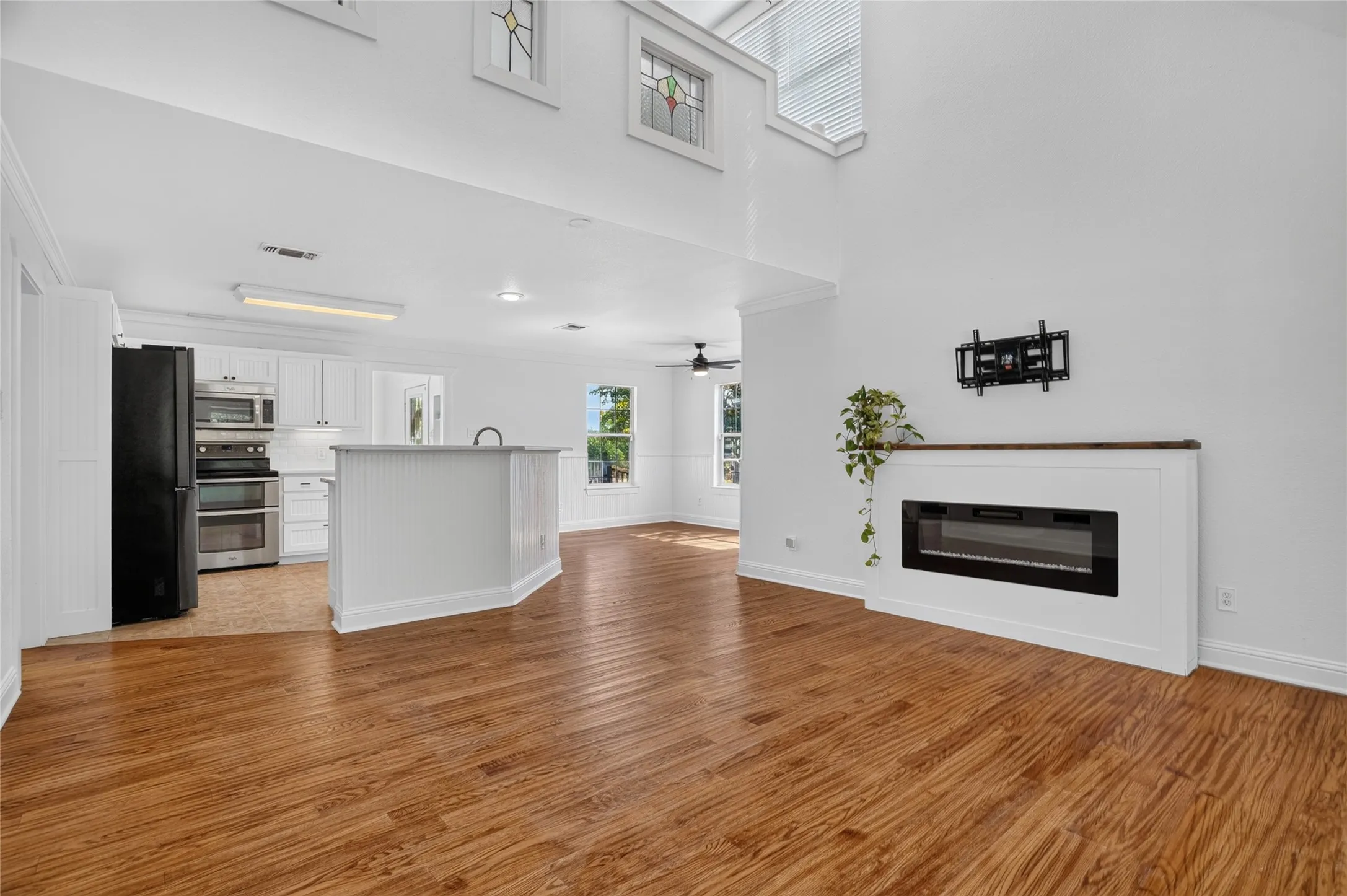 Spacious room with wood-finish flooring, an electric fireplace with a wood mantle, and a white kitchen featuring white cabinetry and black and stainless steel appliances