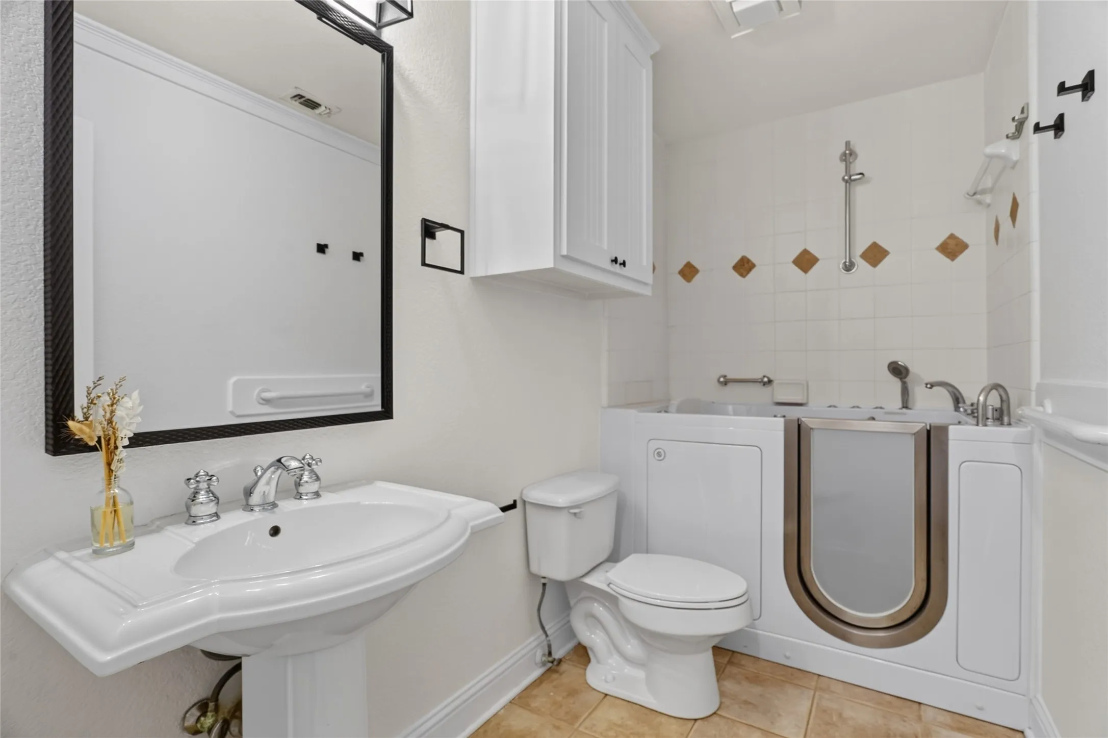 Pedestal sink with chrome fixtures, walk-in bathtub, white wall tiling with decorative accents, and overhead cabinetry