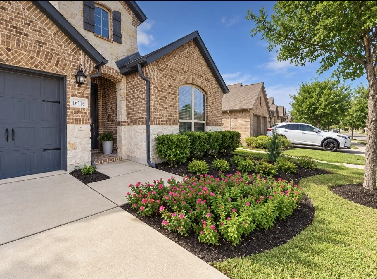 Brick and stone facade, dark gray garage door, arched entry, decorative shutters, and mature landscaping