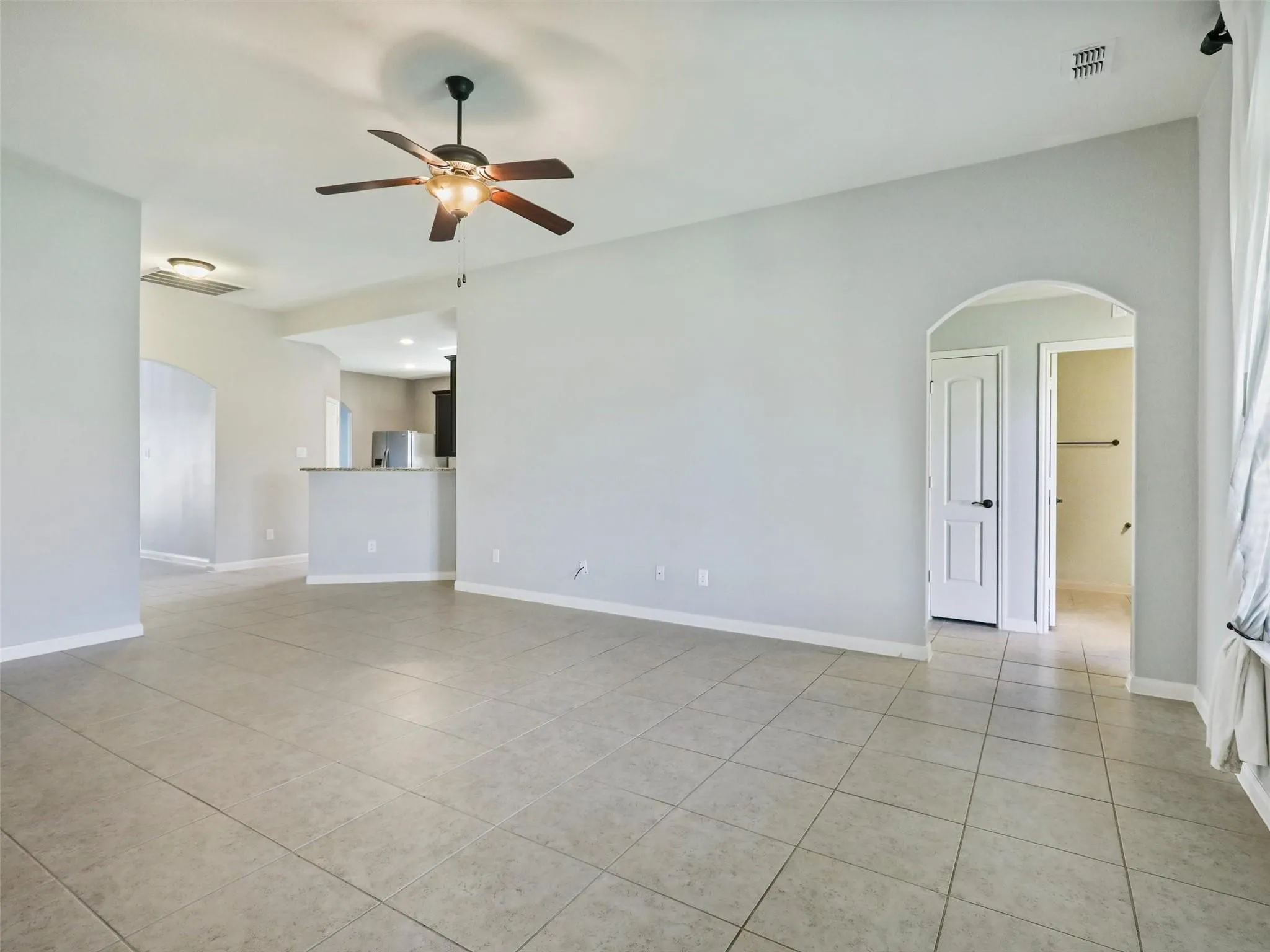 Rectangular tile flooring with a neutral finish, arched doorway, ceiling fan with integrated lighting, and a partial wall separating spaces