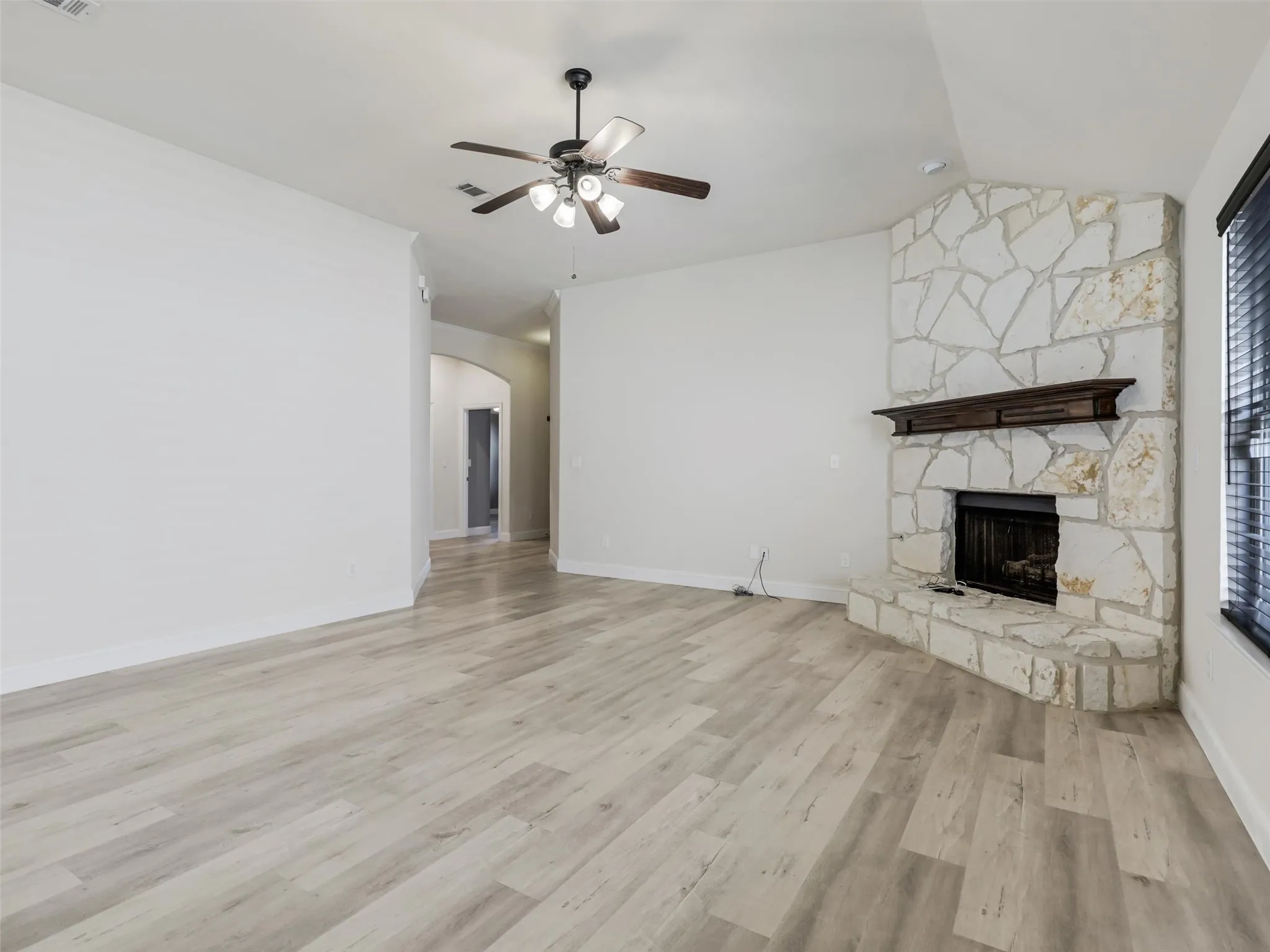 White interior walls with light wood-finish flooring, a corner stone fireplace with a wood mantle, and a ceiling fan with integrated lighting