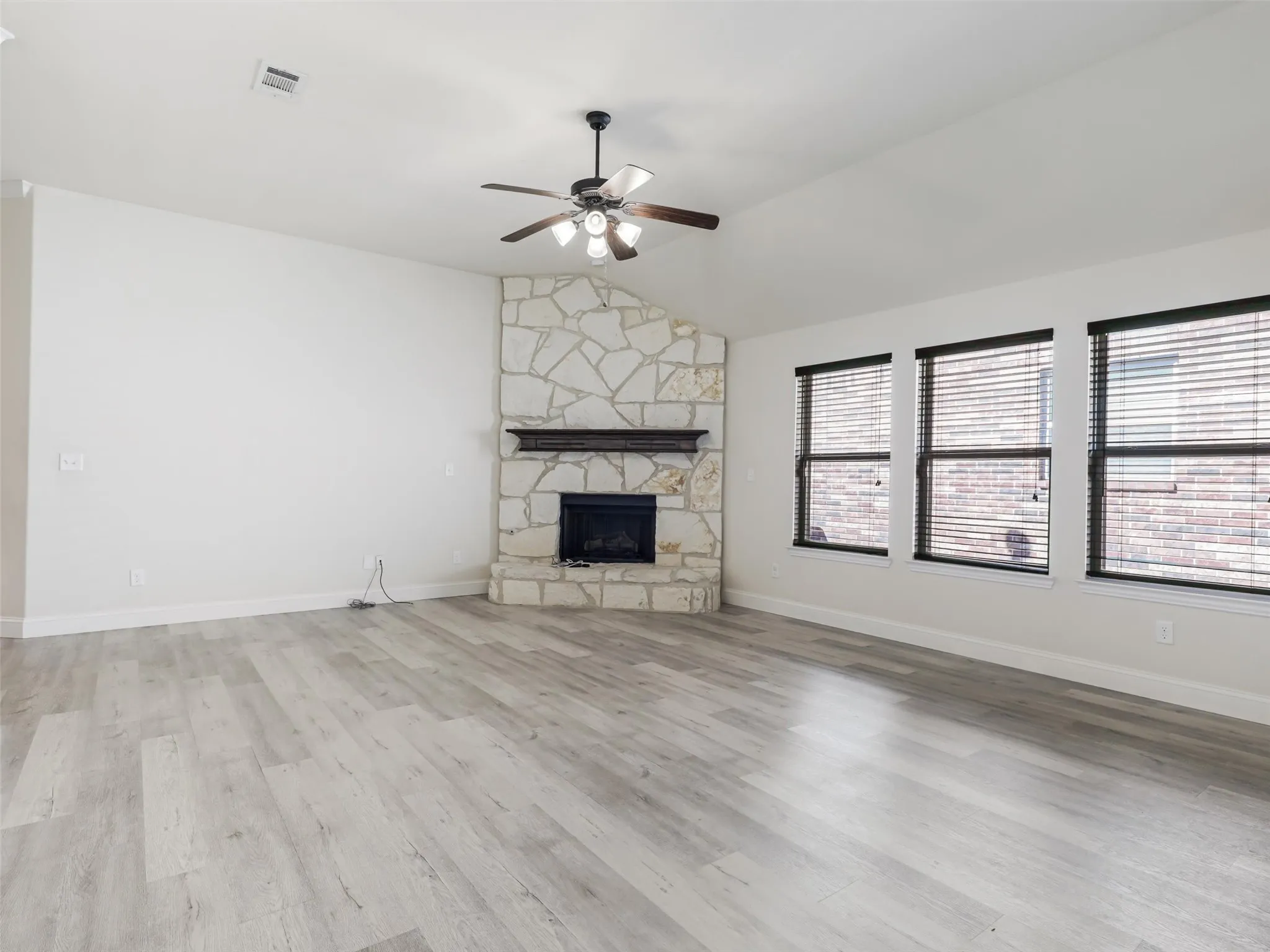 Vaulted ceilings, a stone-clad fireplace with a dark wood mantel, a ceiling fan with integrated lighting, and multiple windows with blinds illuminate the spacious room with wood-finish flooring