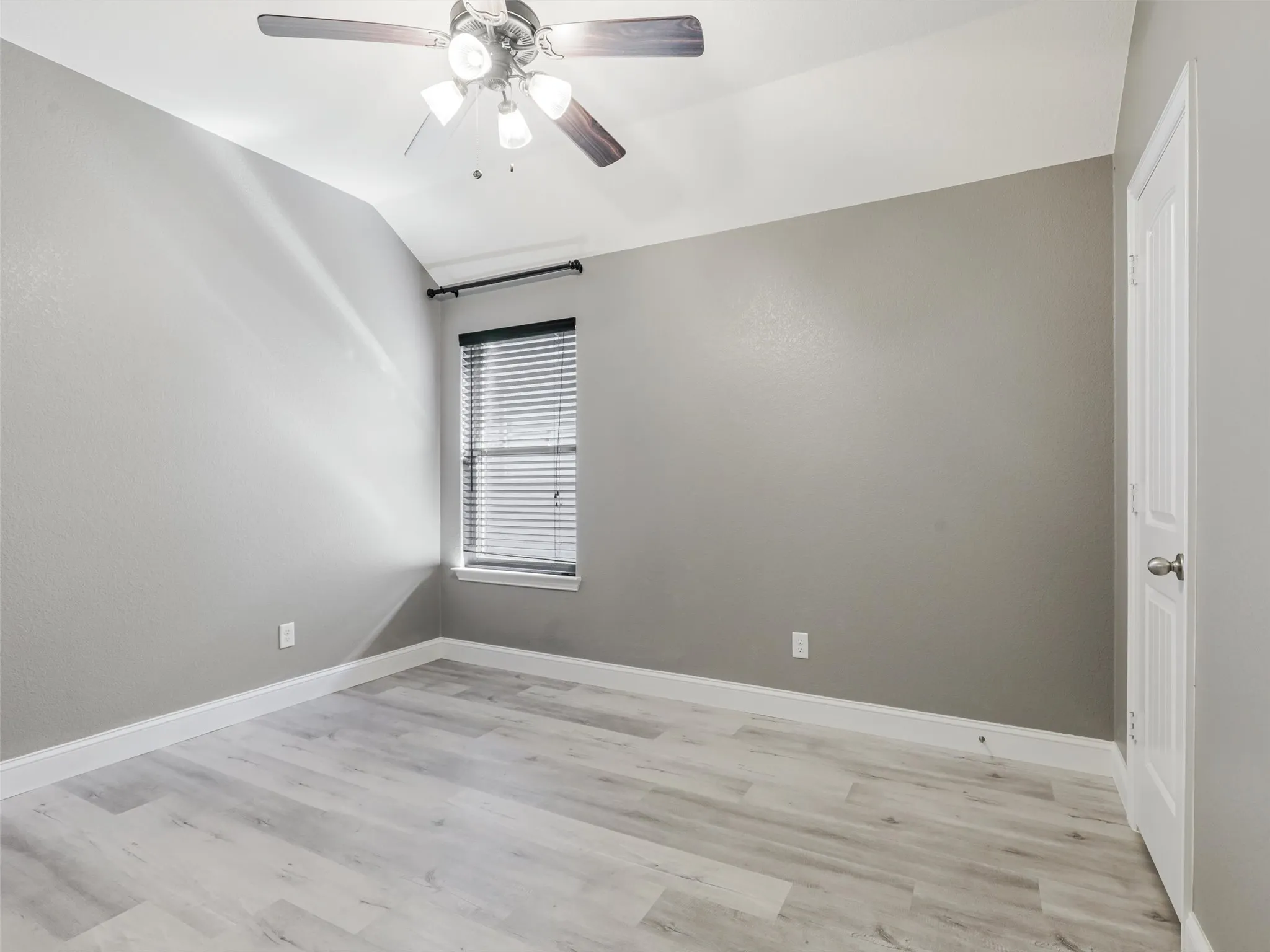 Vaulted ceiling with a ceiling fan, light wood-finish flooring, window with blinds, and white door