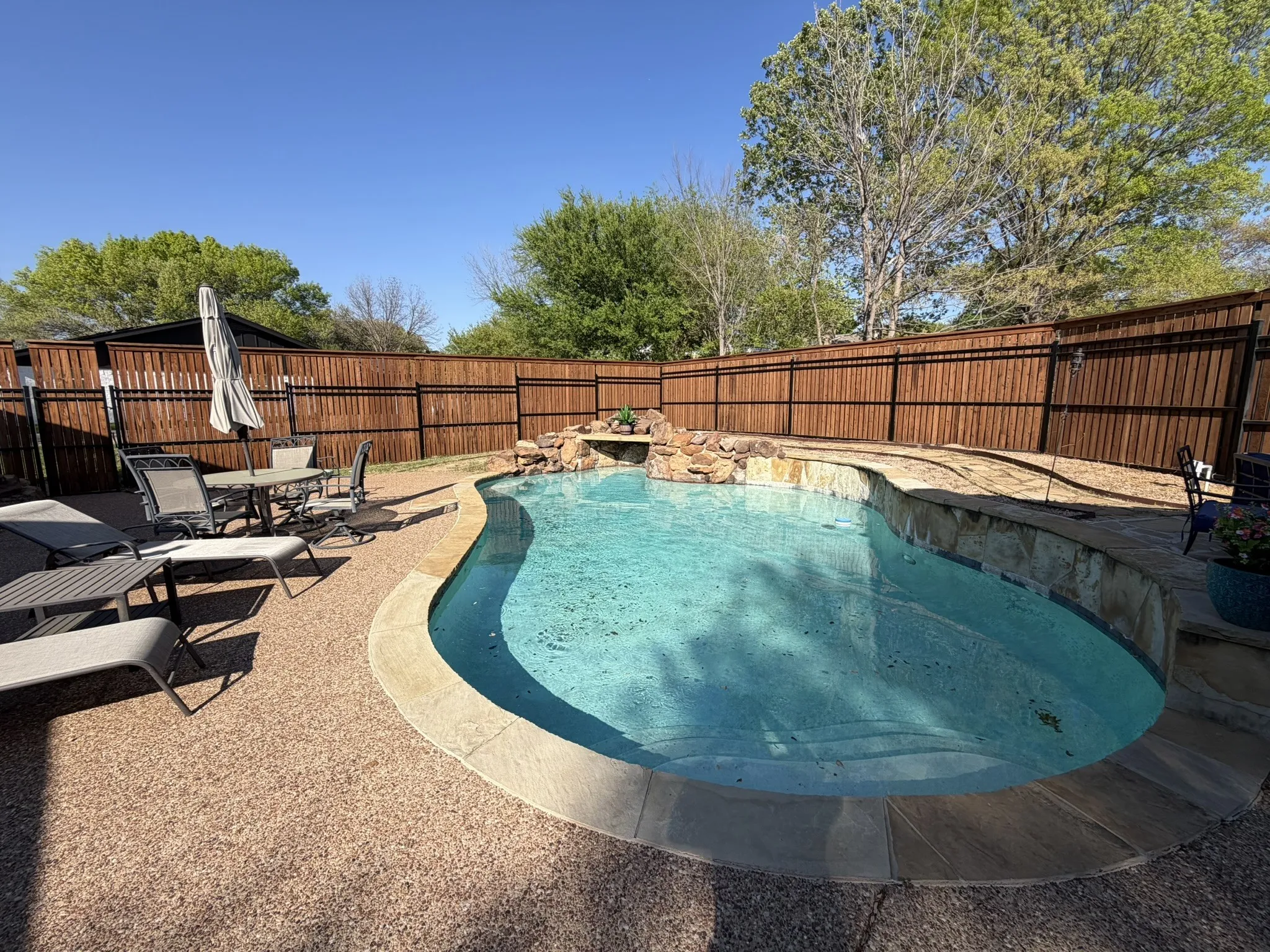 Curved swimming pool with stone coping, a rock waterfall feature, and an adjacent aggregate patio